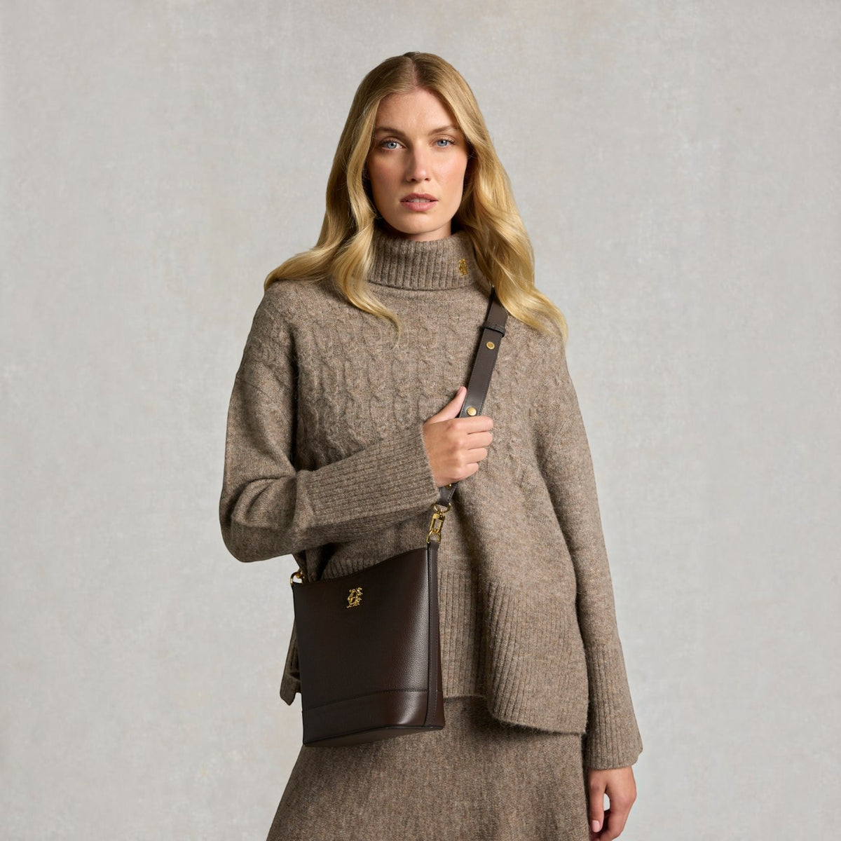 A blonde woman wearing a taupe cable-knit turtleneck sweater and matching skirt, carrying a dark brown leather crossbody bag with gold hardware against a textured light gray background.
