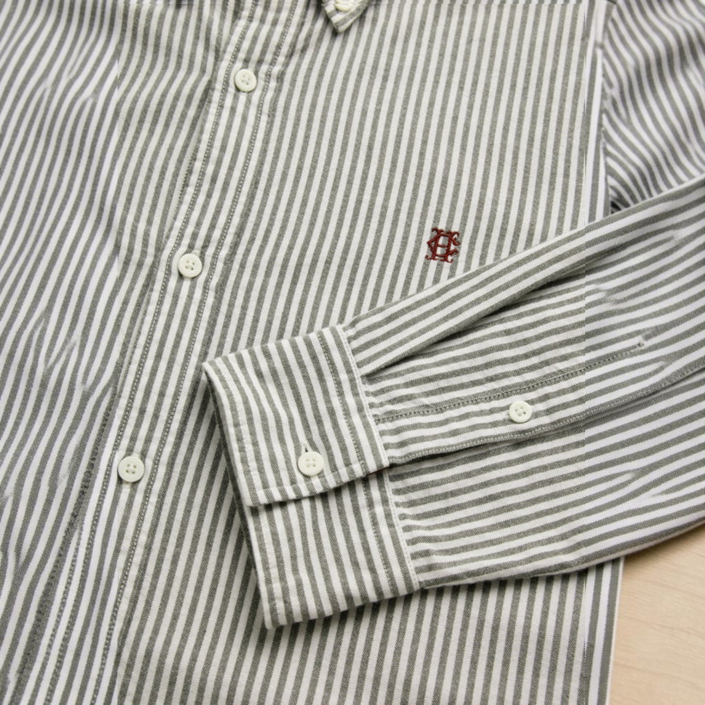 Close-up of a striped long-sleeve button-up shirt with white buttons and a small red embroidered logo on the chest, laid flat on a light wood surface.