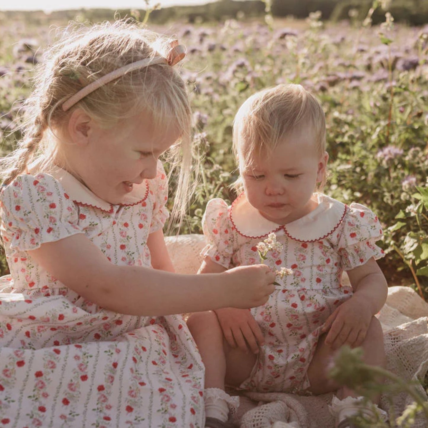 Two young girls in matching floral dresses sitting in a field of flowers.