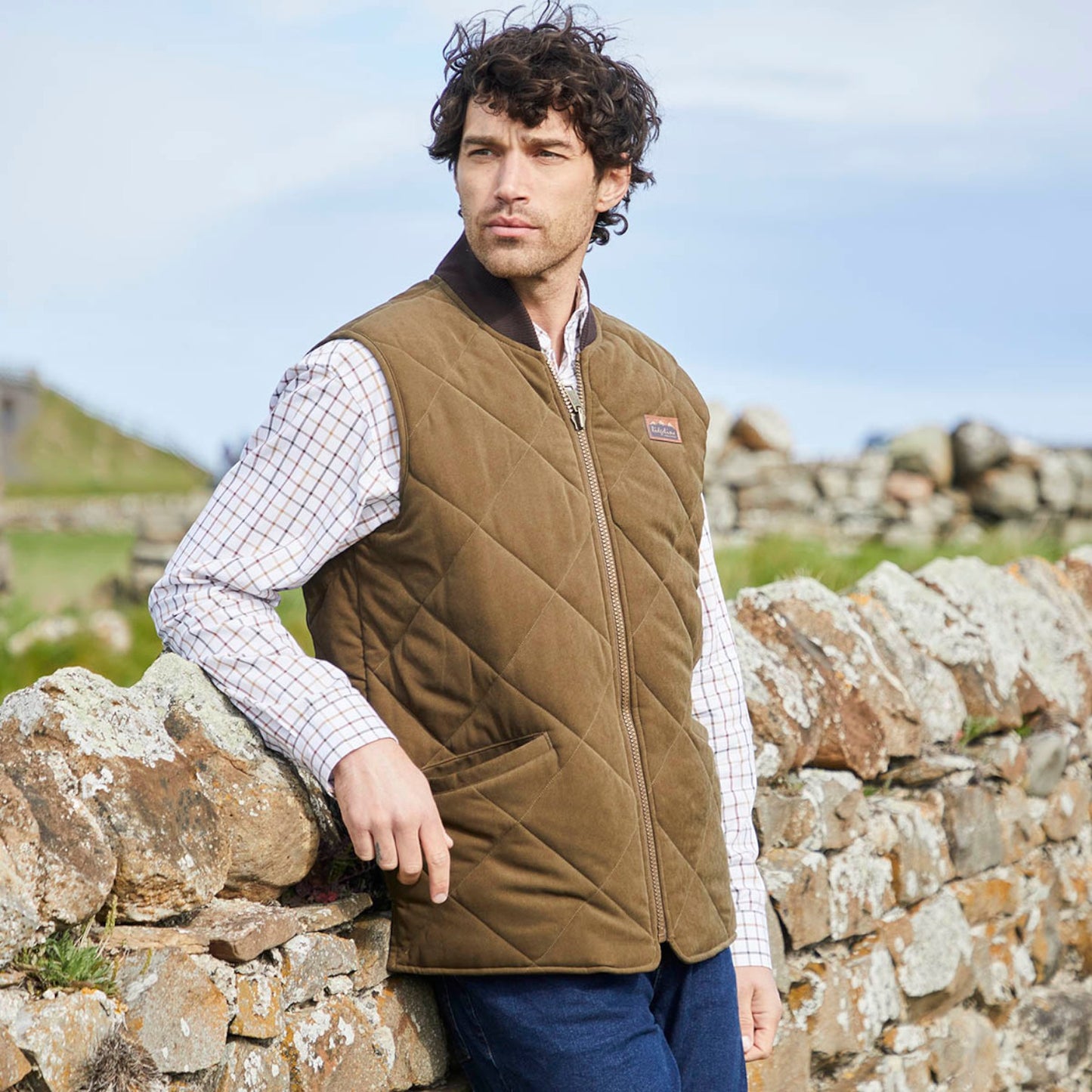 A man wearing a brown diamond-quilted zip-up vest over a white checkered shirt and blue jeans, leaning against a stone wall outdoors.