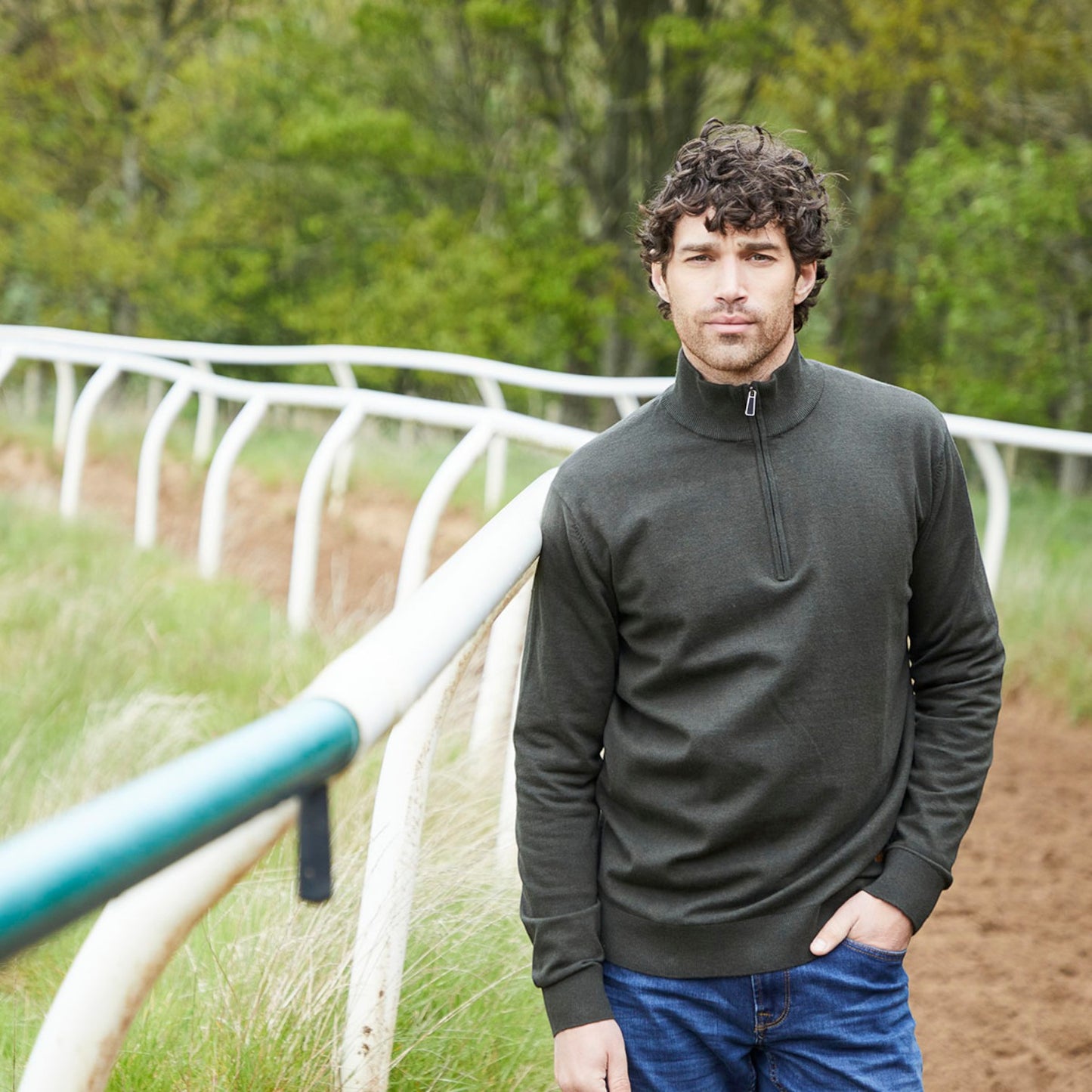 A man wearing a dark olive green quarter-zip long-sleeve sweater and blue jeans, standing outdoors on a dirt track next to a white rail with trees in the background.