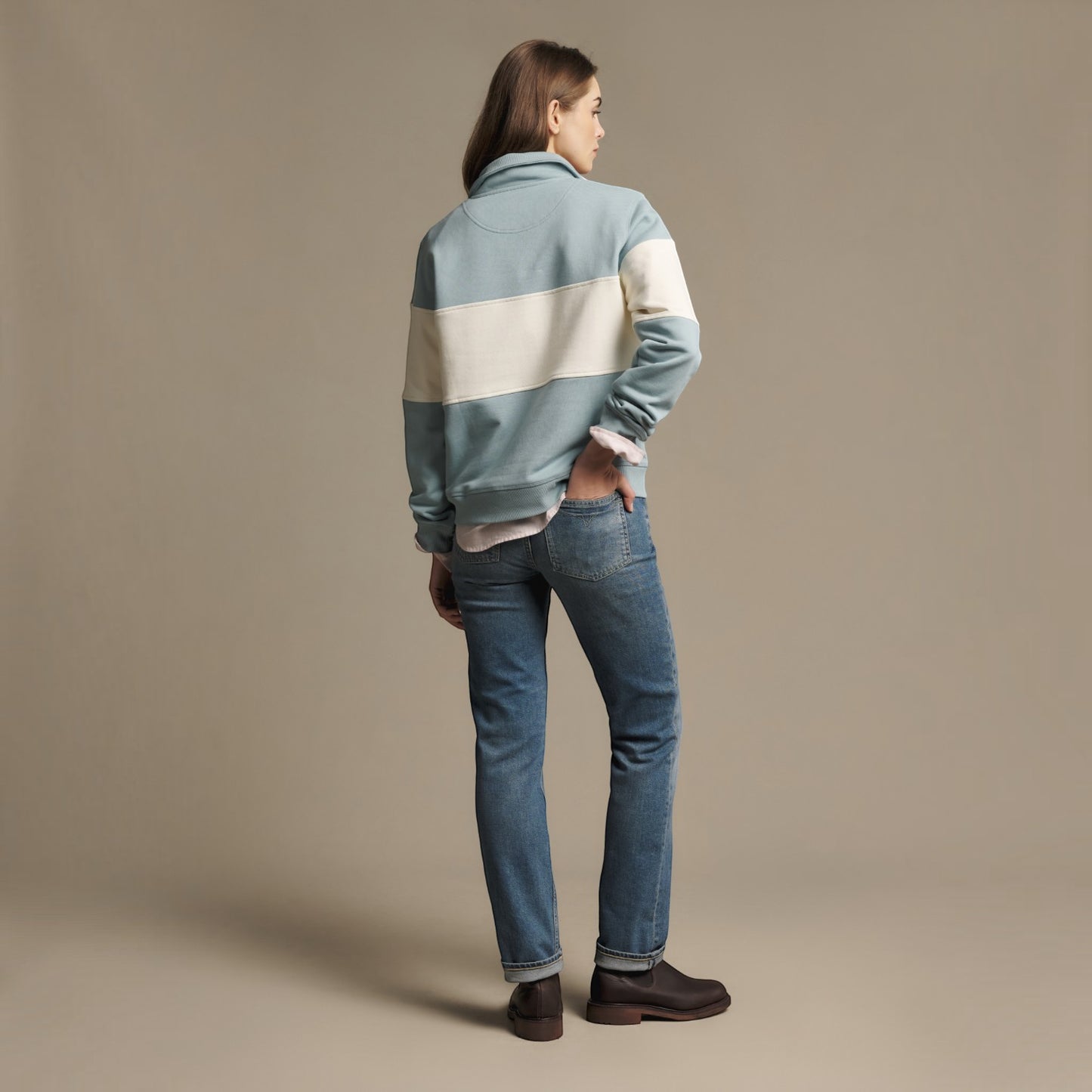 A model shown from the back wearing a light blue and cream color-blocked fleece jacket, blue jeans, and dark brown boots against a solid beige studio background.