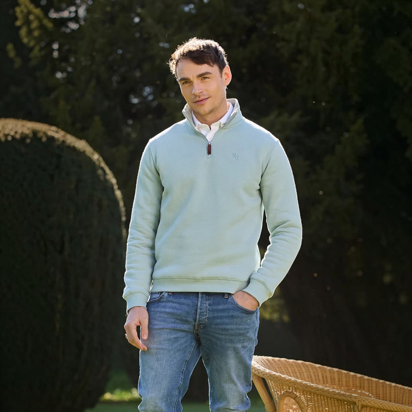Man wearing a light blue sweater and blue jeans standing outdoors with trees in the background