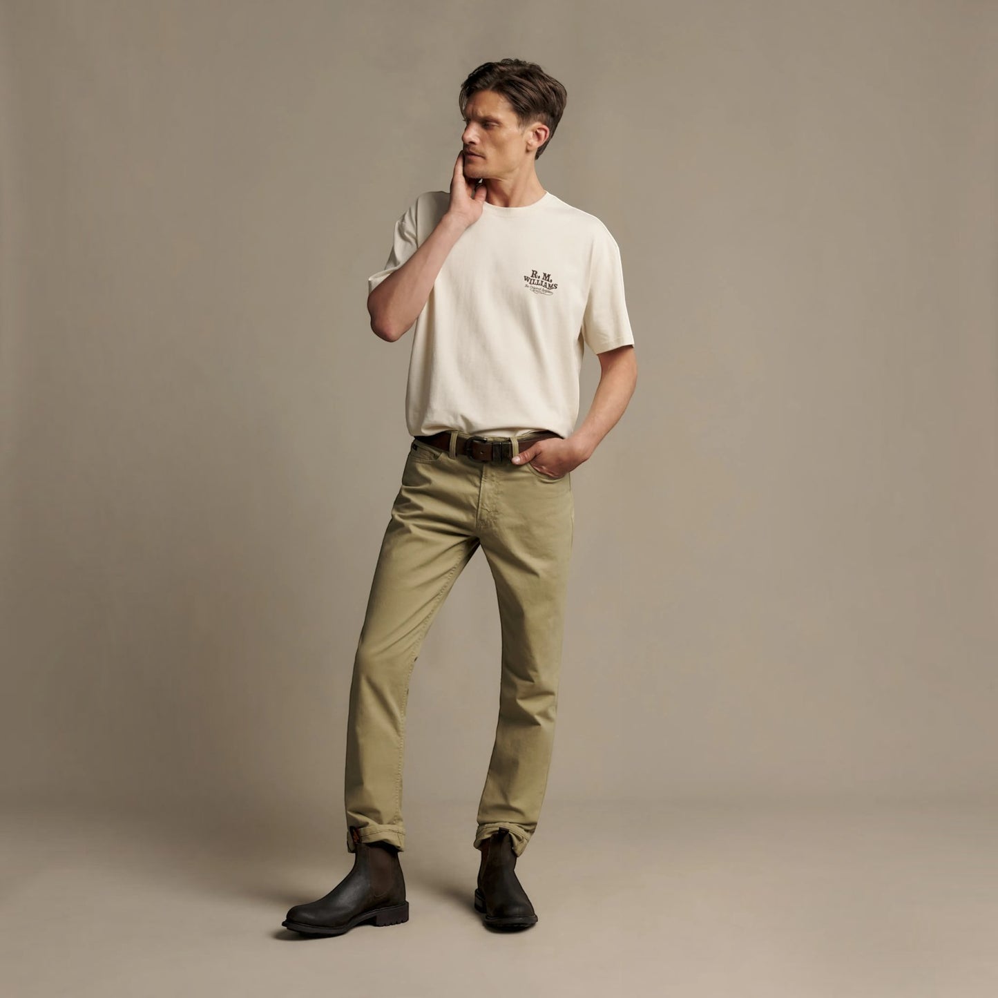 A man modeling a cream short-sleeve crewneck t-shirt with a small black chest logo, paired with olive green trousers and black boots, standing against a solid beige studio backdrop.
