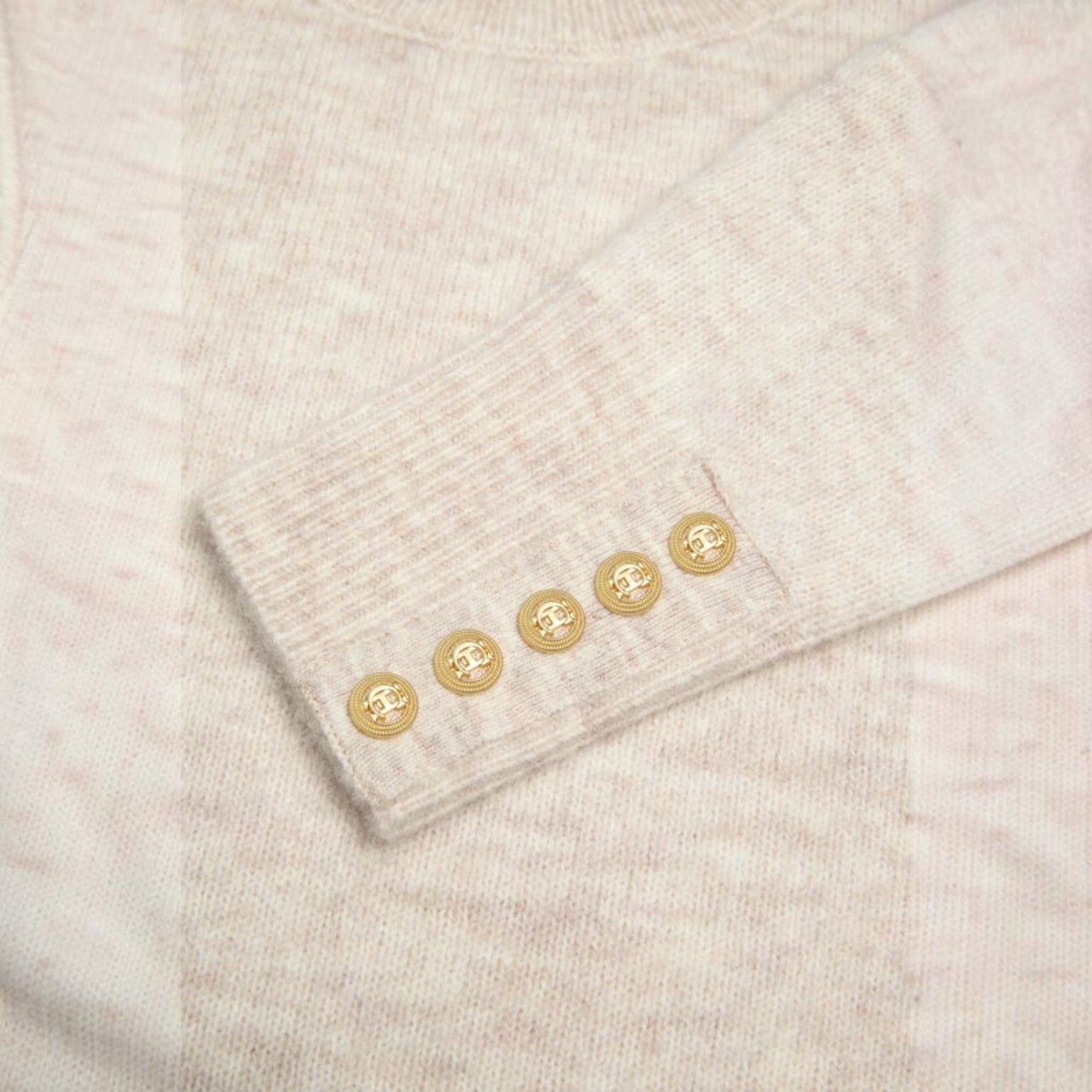 Close-up of a light beige knit sweater sleeve with five decorative gold buttons on the cuff.