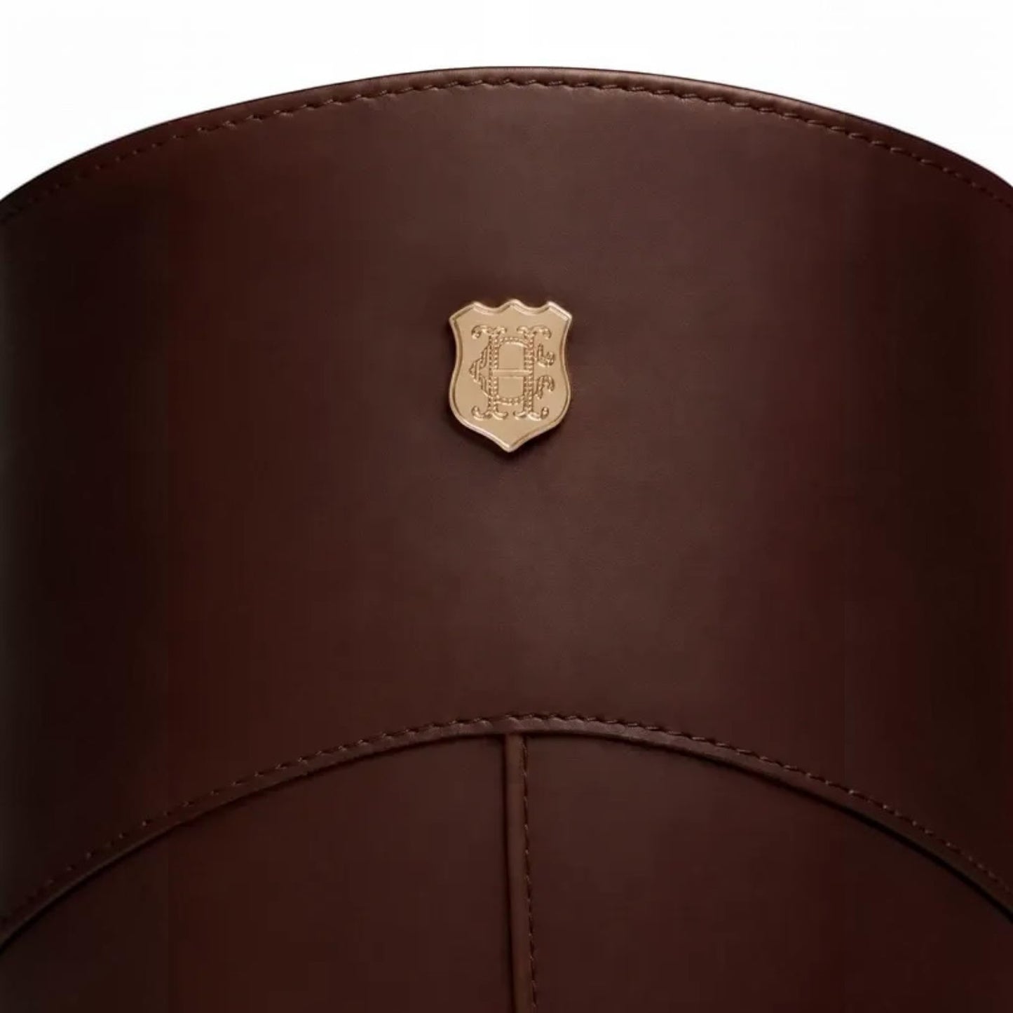 Close-up of a dark brown leather cylindrical product, likely a bag or case, featuring a small gold shield-shaped metal emblem on the front.