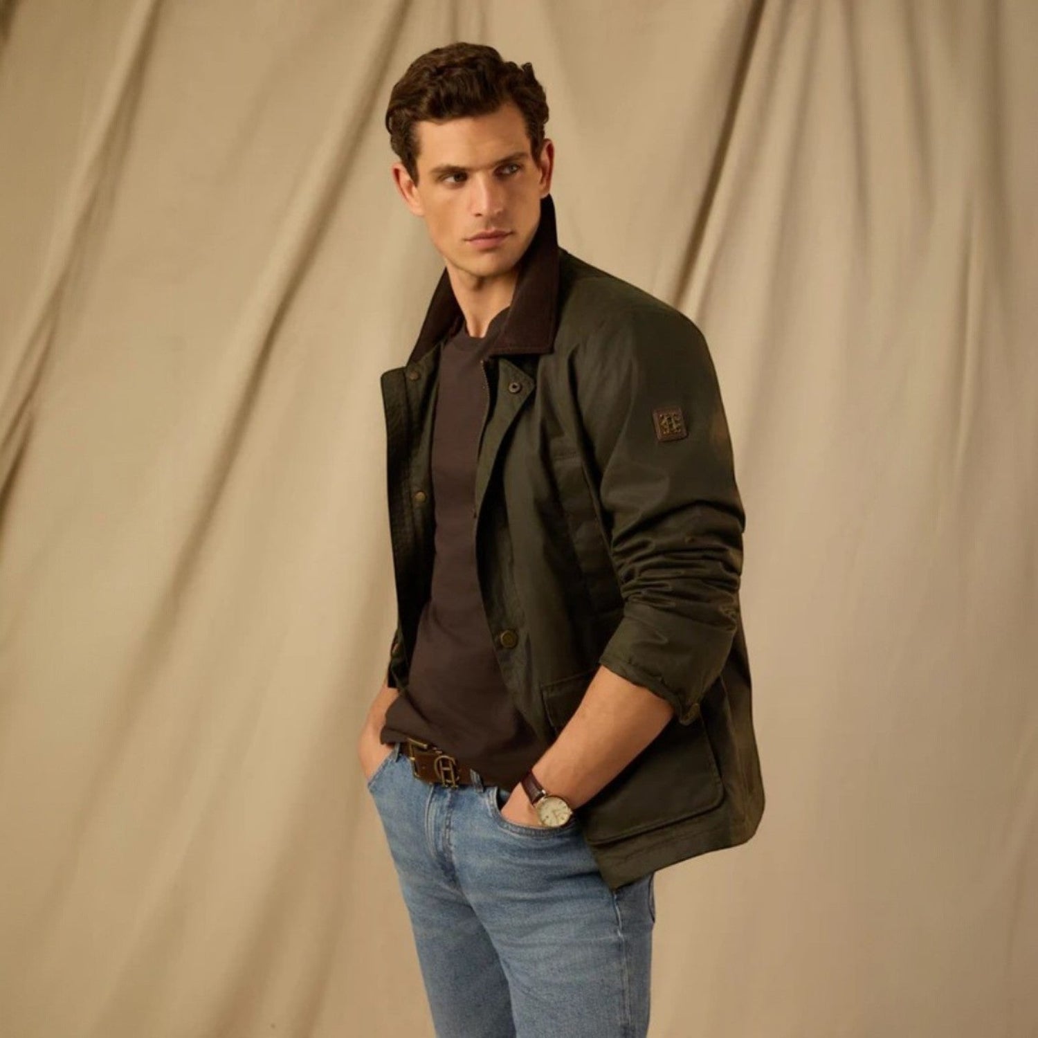 Man wearing a dark green jacket and blue jeans against a beige background