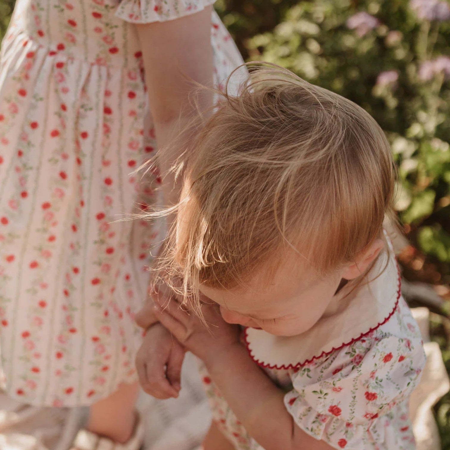 Two children in matching floral dresses standing outdoors with greenery in the background.