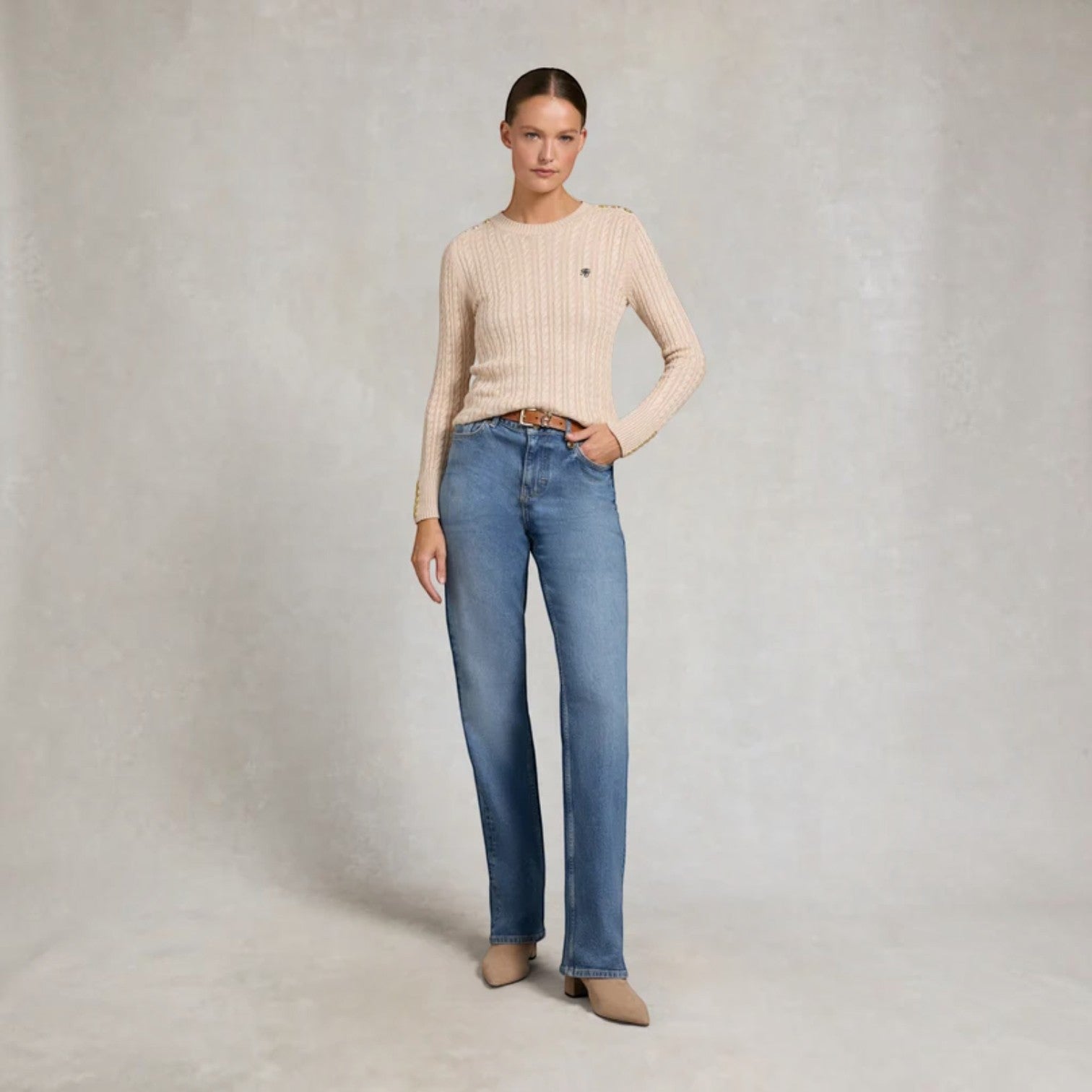 A woman stands against a textured light gray studio backdrop wearing a beige ribbed knit sweater, blue jeans, and beige pointed shoes.