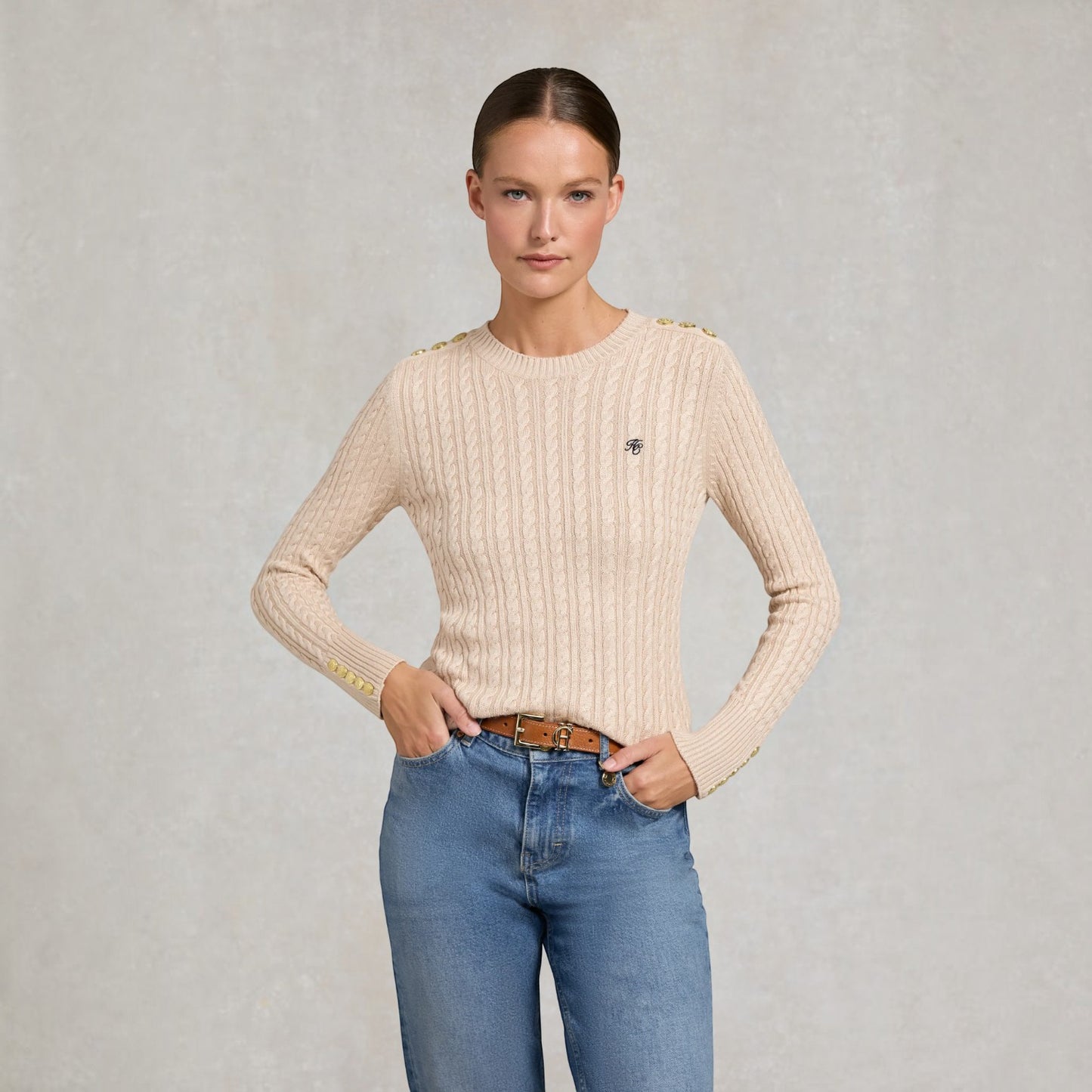A woman modeling a beige cable-knit sweater with gold button accents at the shoulders and a small black embroidered logo on the chest, paired with blue jeans against a textured gray background.