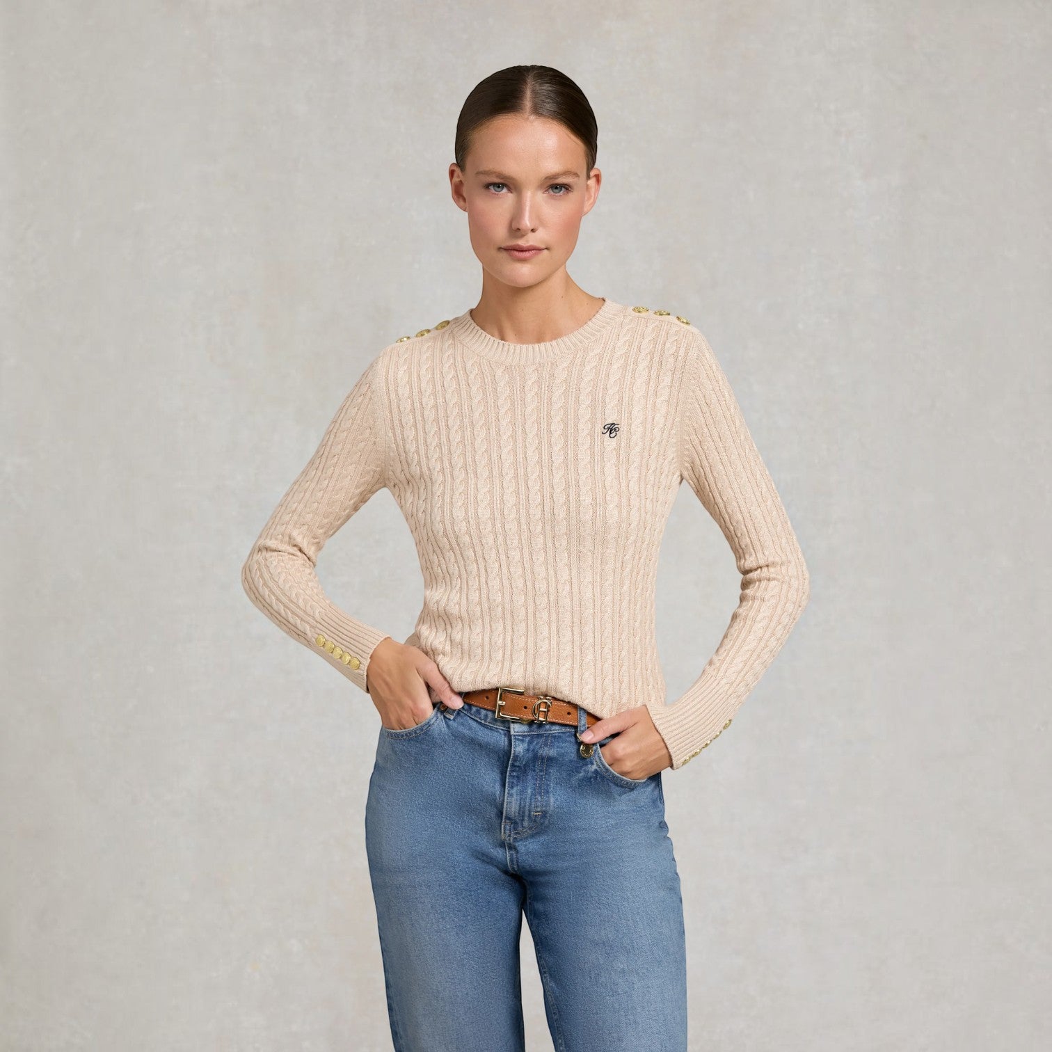 A woman modeling a beige cable-knit sweater with gold button accents at the shoulders and a small black embroidered logo on the chest, paired with blue jeans against a textured gray background.