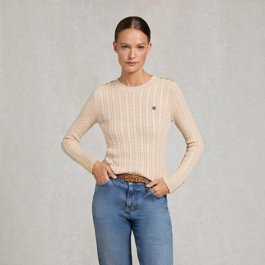 A woman modeling a beige cable-knit sweater with gold button accents at the shoulders and a small black embroidered logo on the chest, paired with blue jeans against a textured gray background.