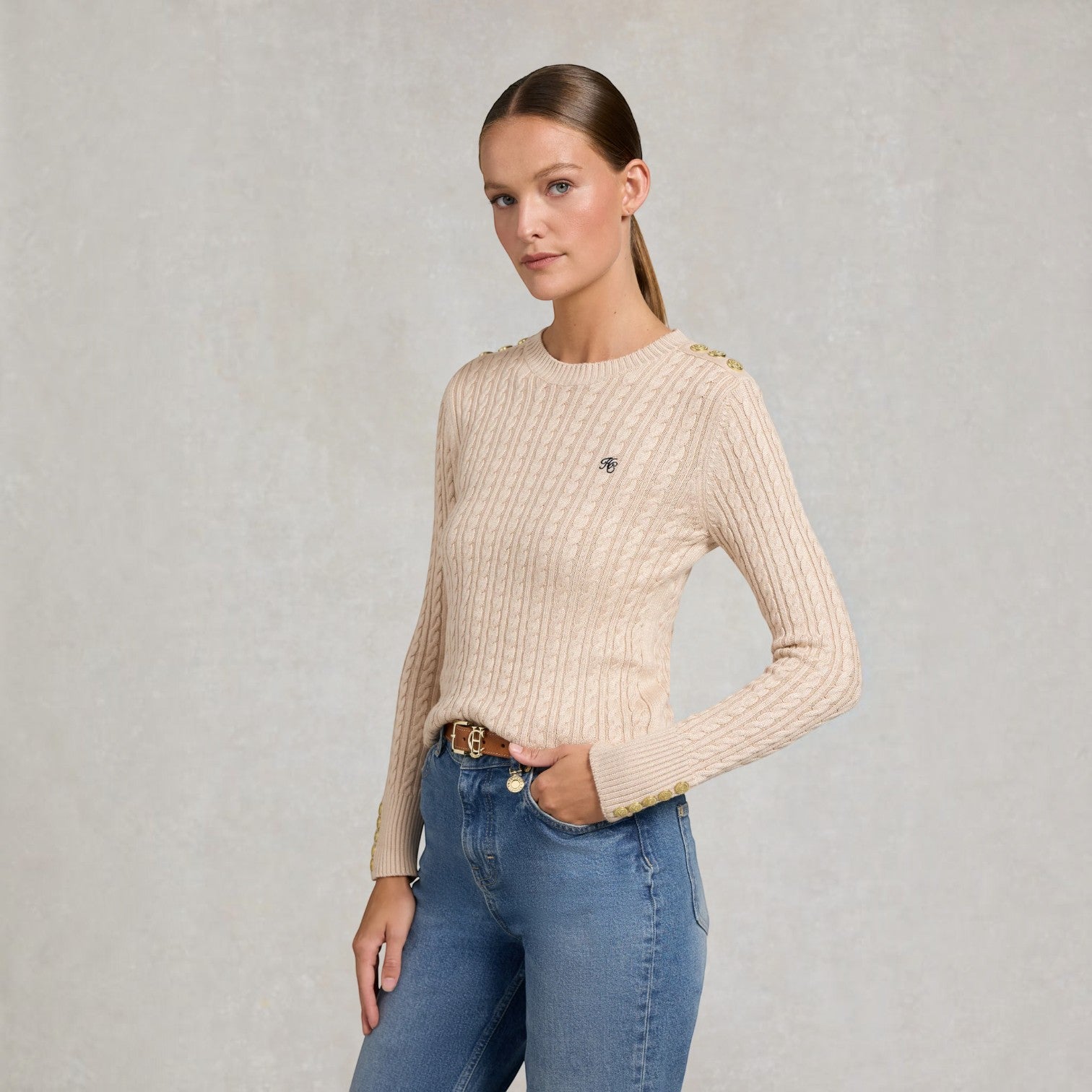 A woman modeling a beige cable-knit crewneck sweater with gold button accents on the shoulders and a small embroidered logo on the chest, paired with blue jeans against a textured light gray background.