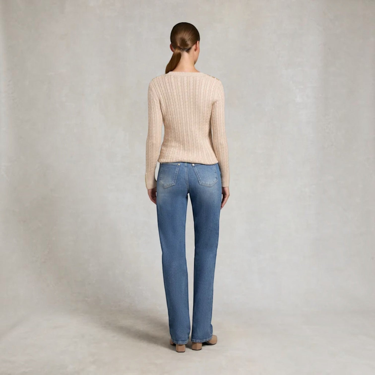 A woman viewed from behind wearing a beige ribbed knit long-sleeve top and blue jeans, standing against a textured light gray studio backdrop.