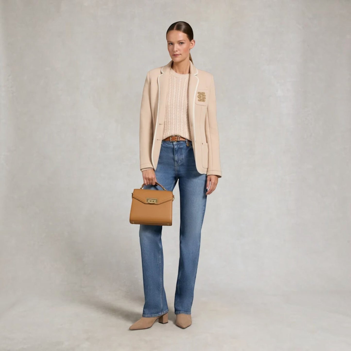A woman stands against a textured light gray studio backdrop wearing a beige blazer over a ribbed cream top, blue jeans, and beige pointed shoes, while holding a tan structured handbag.