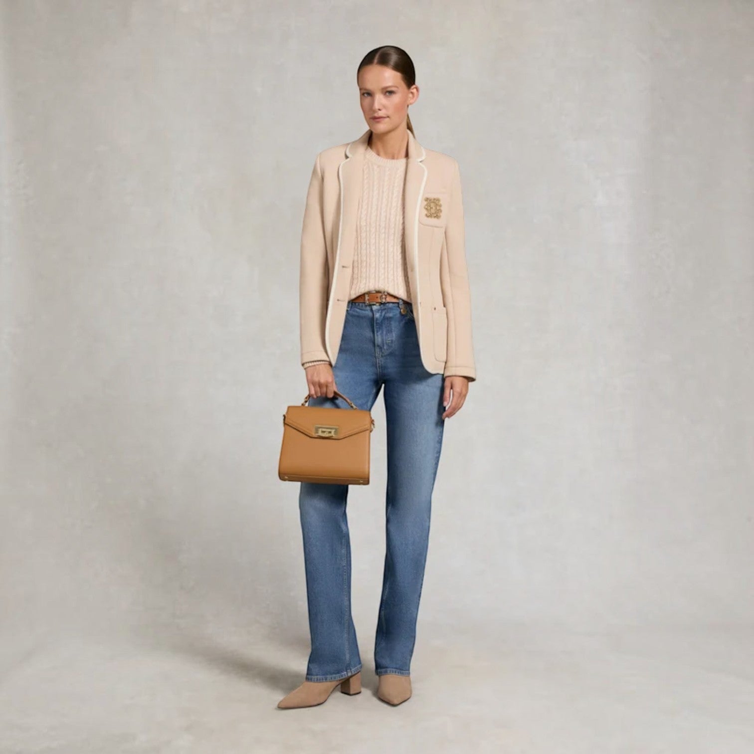 A woman stands against a textured light gray studio backdrop wearing a beige blazer over a ribbed cream top, blue jeans, and beige pointed shoes, while holding a tan structured handbag.