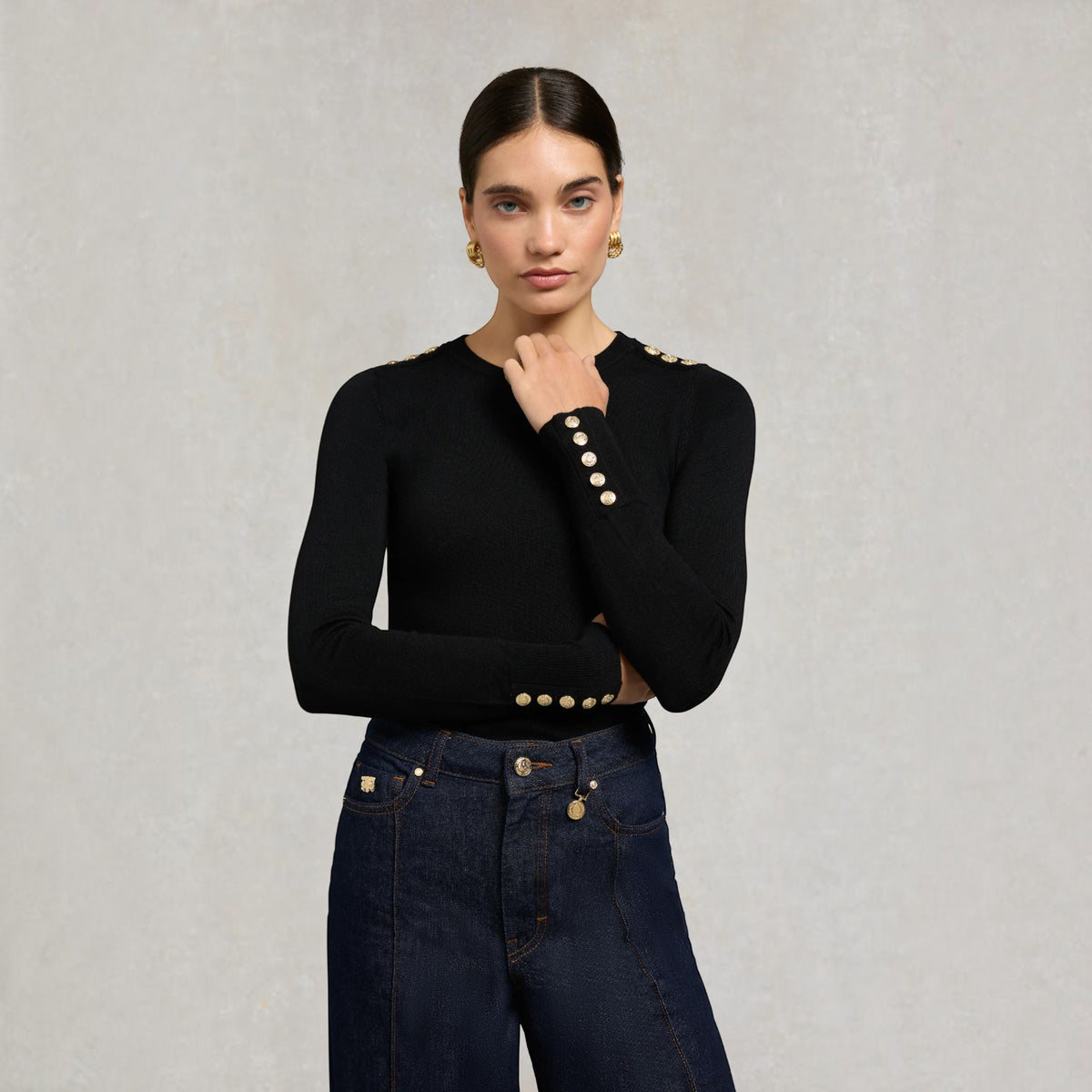 A woman modeling a black long-sleeve knit top with decorative gold-tone buttons on the shoulders and cuffs, paired with dark blue jeans, against a textured light gray background.