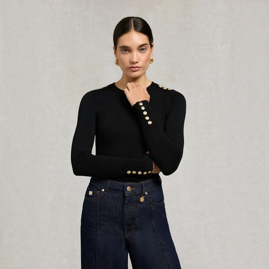 A woman modeling a black long-sleeve knit top with decorative gold-tone buttons on the shoulders and cuffs, paired with dark blue jeans, against a textured light gray background.