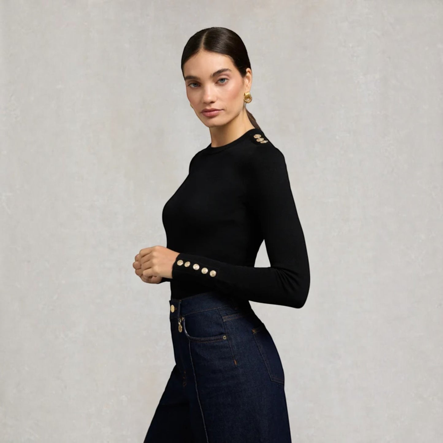 A woman modeling a fitted black long-sleeve top with decorative gold buttons on the shoulders and cuffs, paired with dark blue jeans against a textured light gray background.
