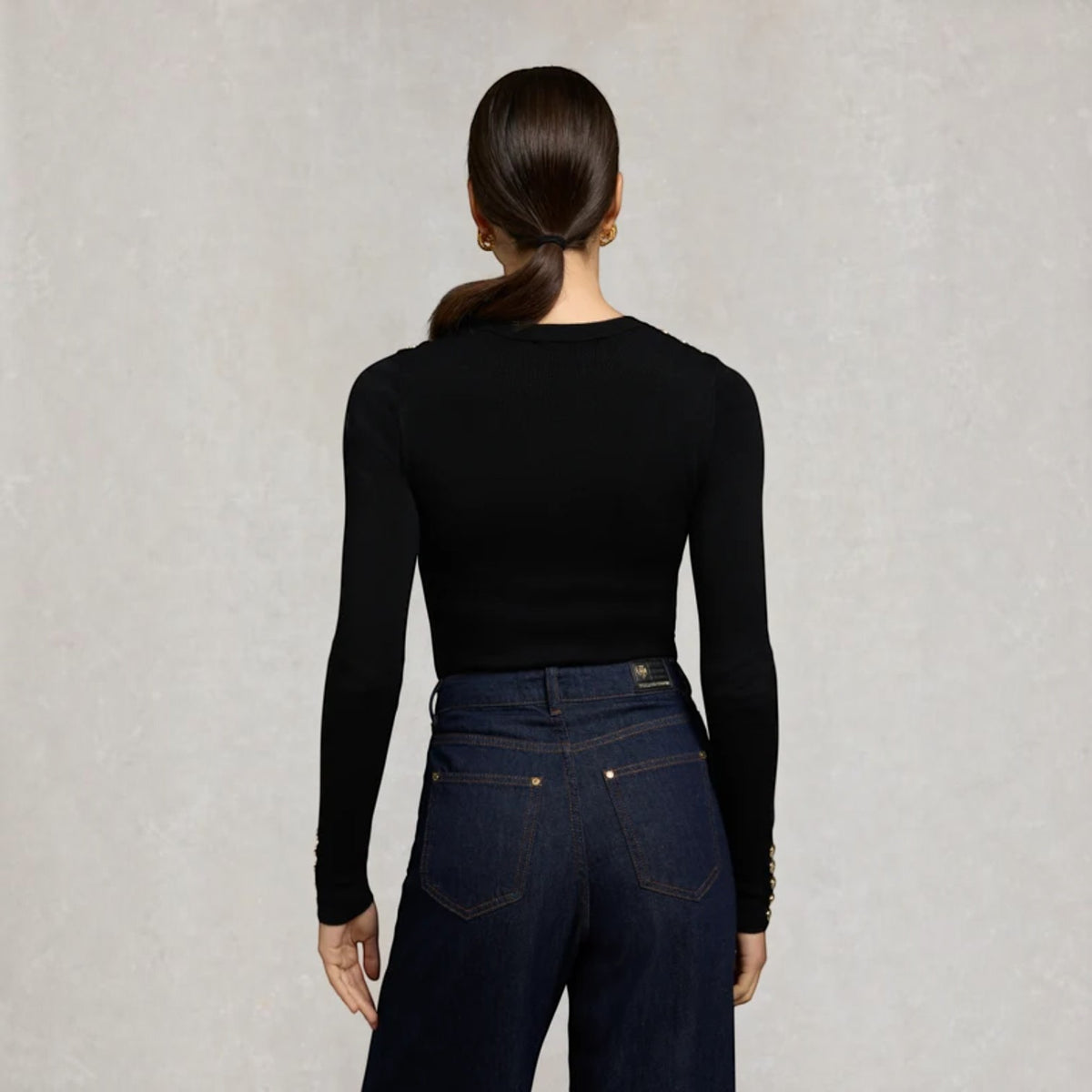 A woman shown from the back wearing a fitted black long-sleeve top and dark blue jeans against a light textured gray background.