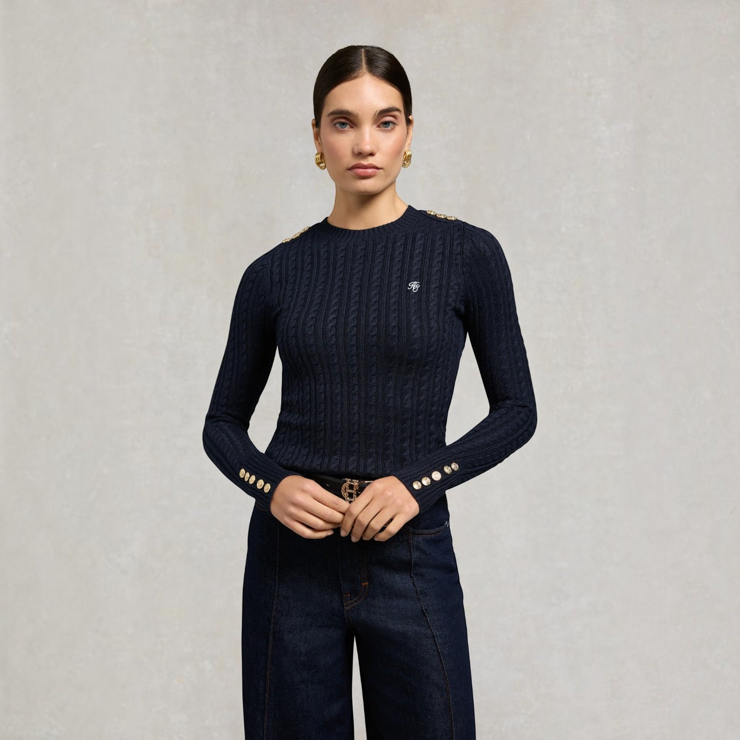 A woman stands against a light grey textured wall wearing a dark navy cable-knit long-sleeve top with gold button accents on the cuffs and shoulders, paired with dark blue jeans.