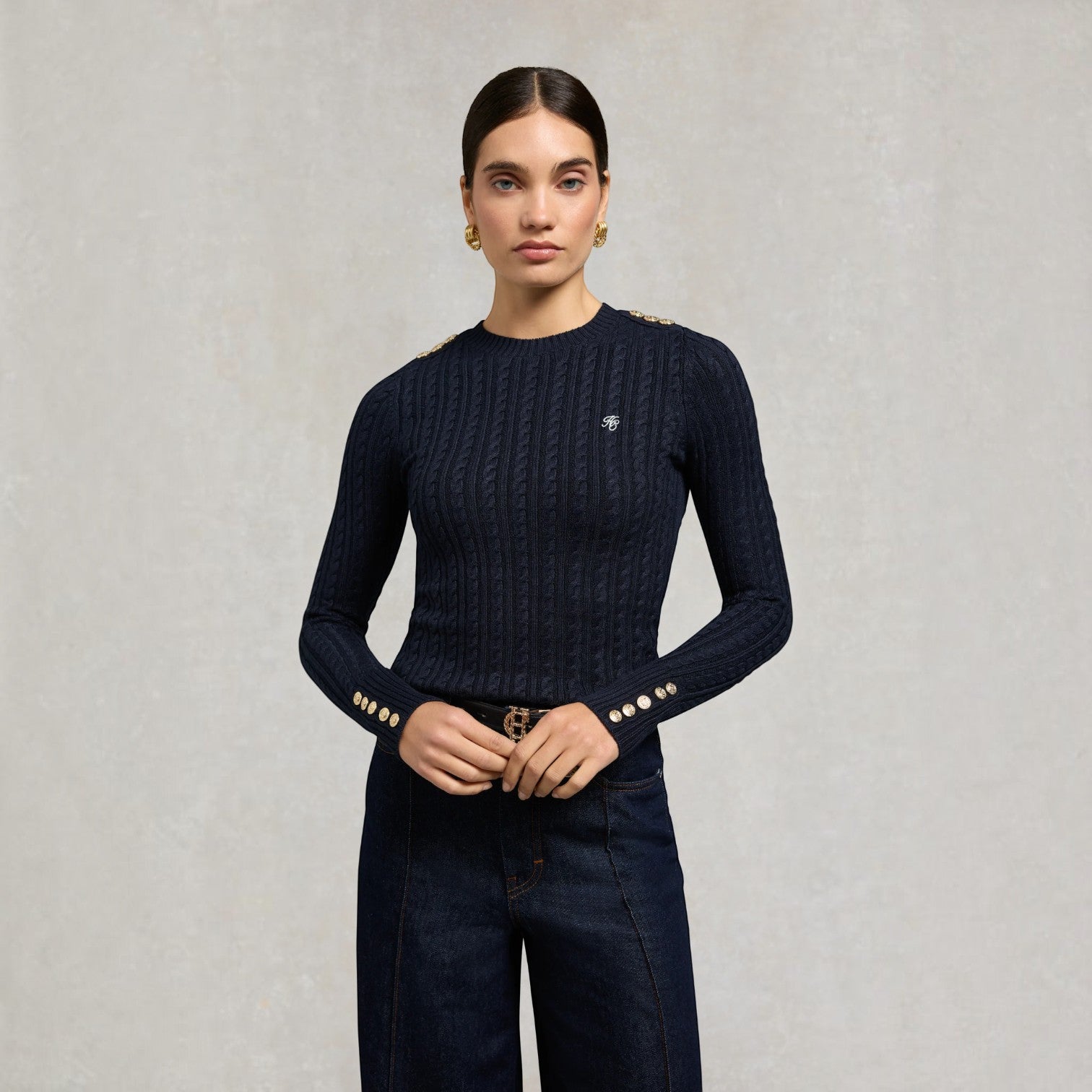A woman stands against a light grey textured wall wearing a dark navy cable-knit long-sleeve top with gold button accents on the cuffs and shoulders, paired with dark blue jeans.