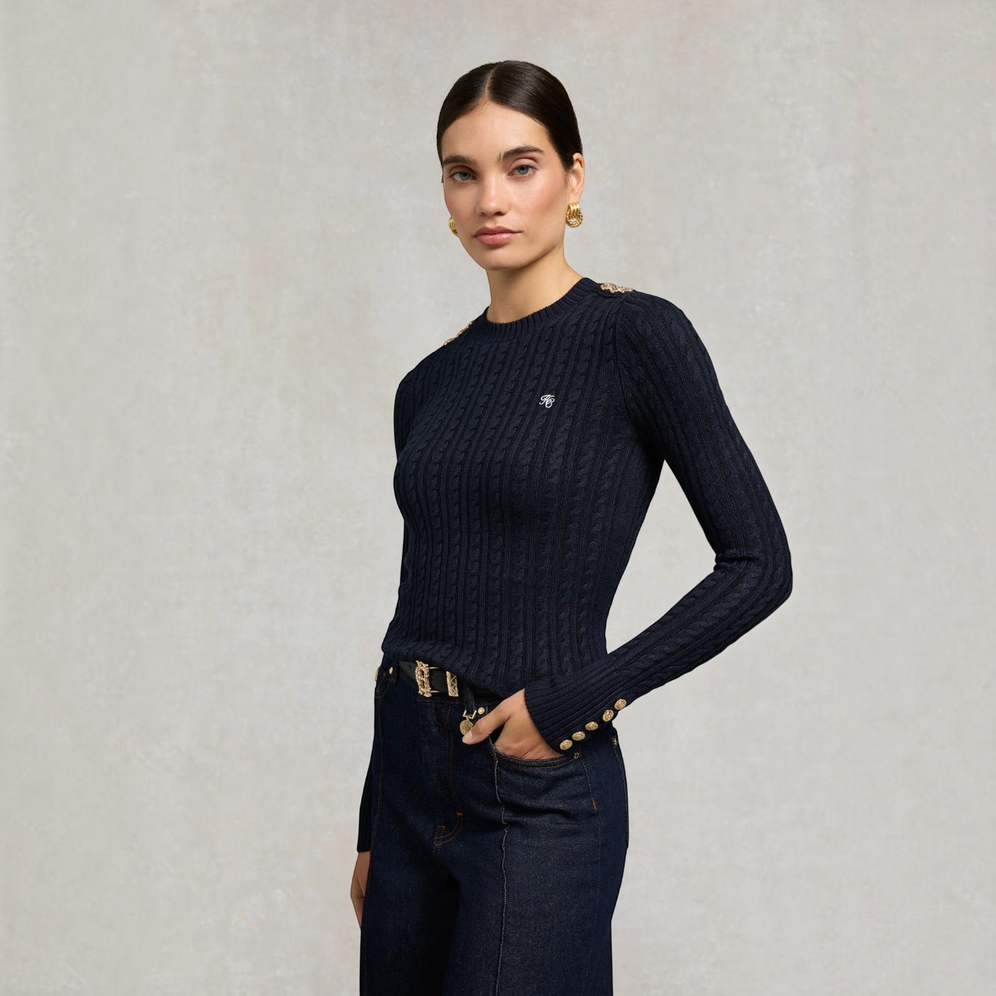 A woman modeling a dark navy blue cable-knit long-sleeve sweater with gold button accents on the cuffs and shoulders, paired with dark jeans and gold earrings, against a textured light gray background.