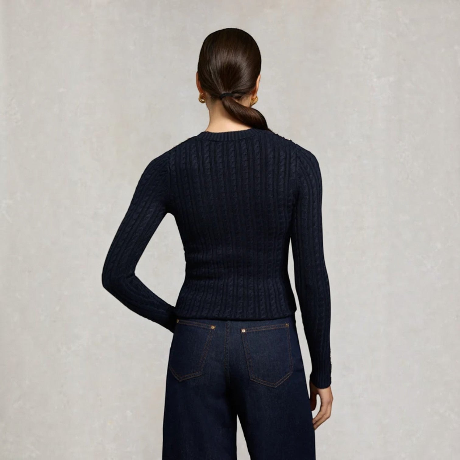 A woman shown from the back wearing a dark navy ribbed long-sleeve top and dark blue jeans against a textured light gray background.