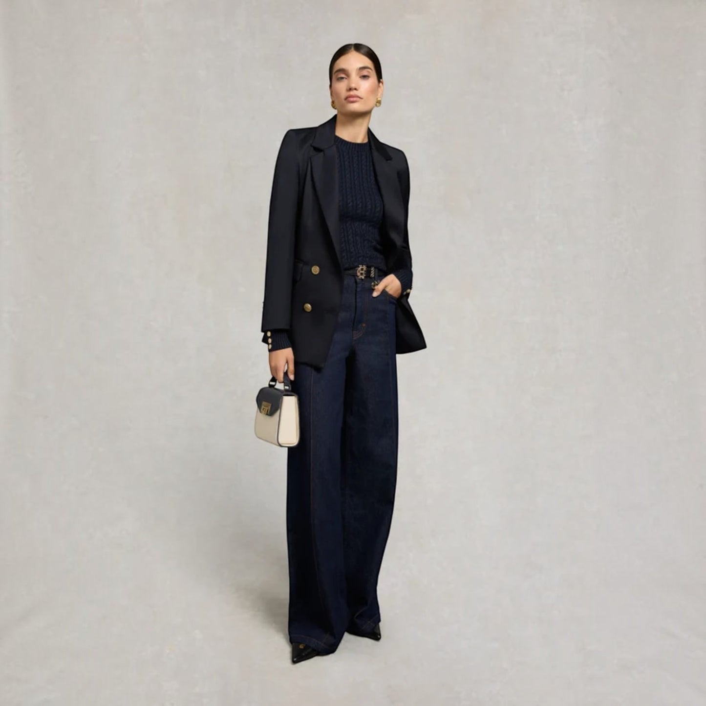 A model stands against a textured light gray studio background wearing a dark navy double-breasted blazer, matching wide-leg trousers, and a ribbed navy top. She holds a small cream and navy handbag.