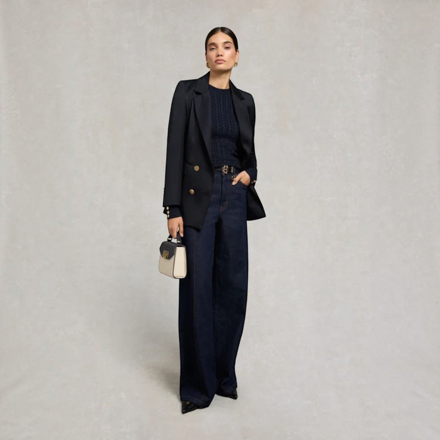 A model stands against a textured light gray studio background wearing a dark navy double-breasted blazer, matching wide-leg trousers, and a ribbed navy top. She holds a small cream and navy handbag.