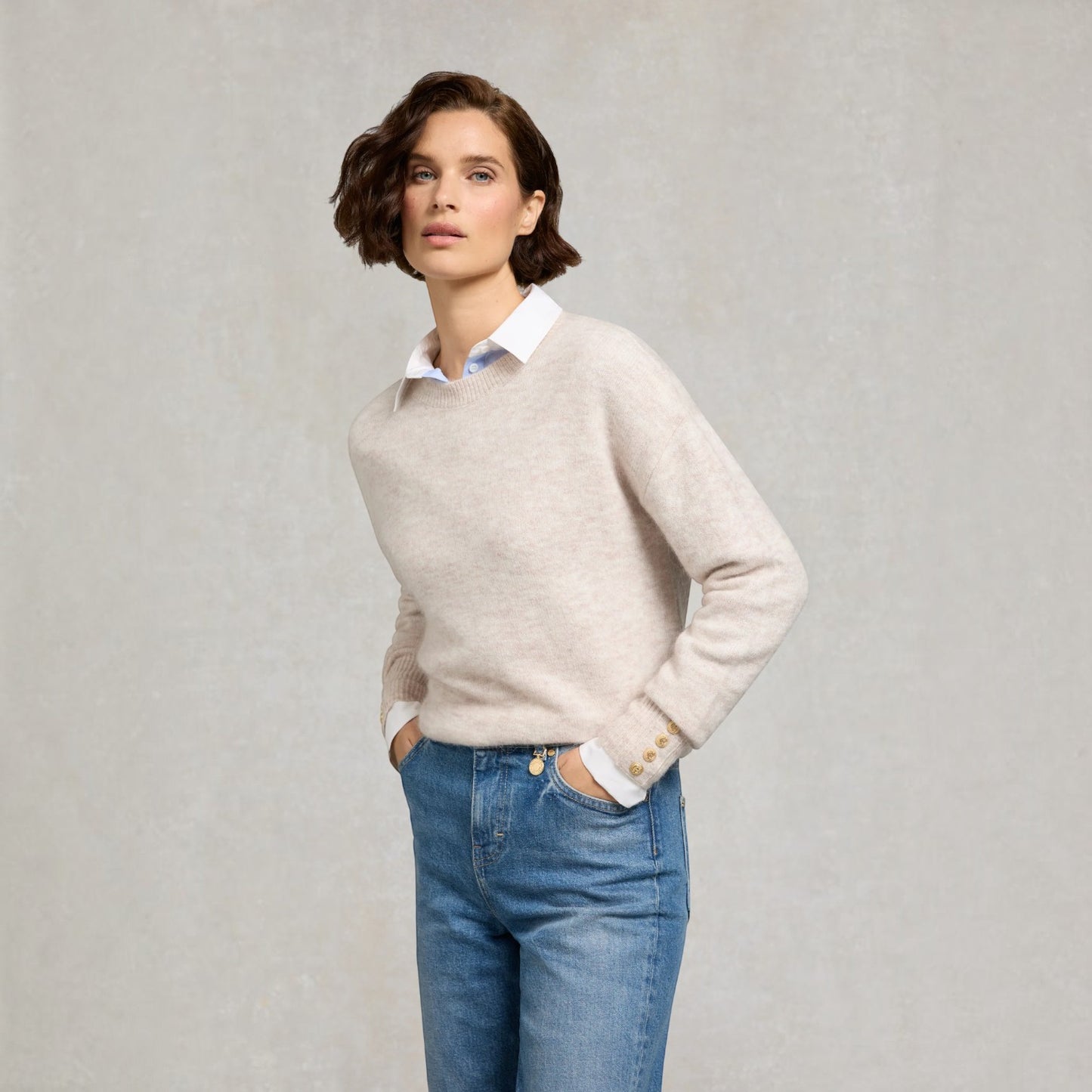 A woman modeling a light beige knit sweater over a white collared shirt and blue jeans, standing against a textured light gray studio background.