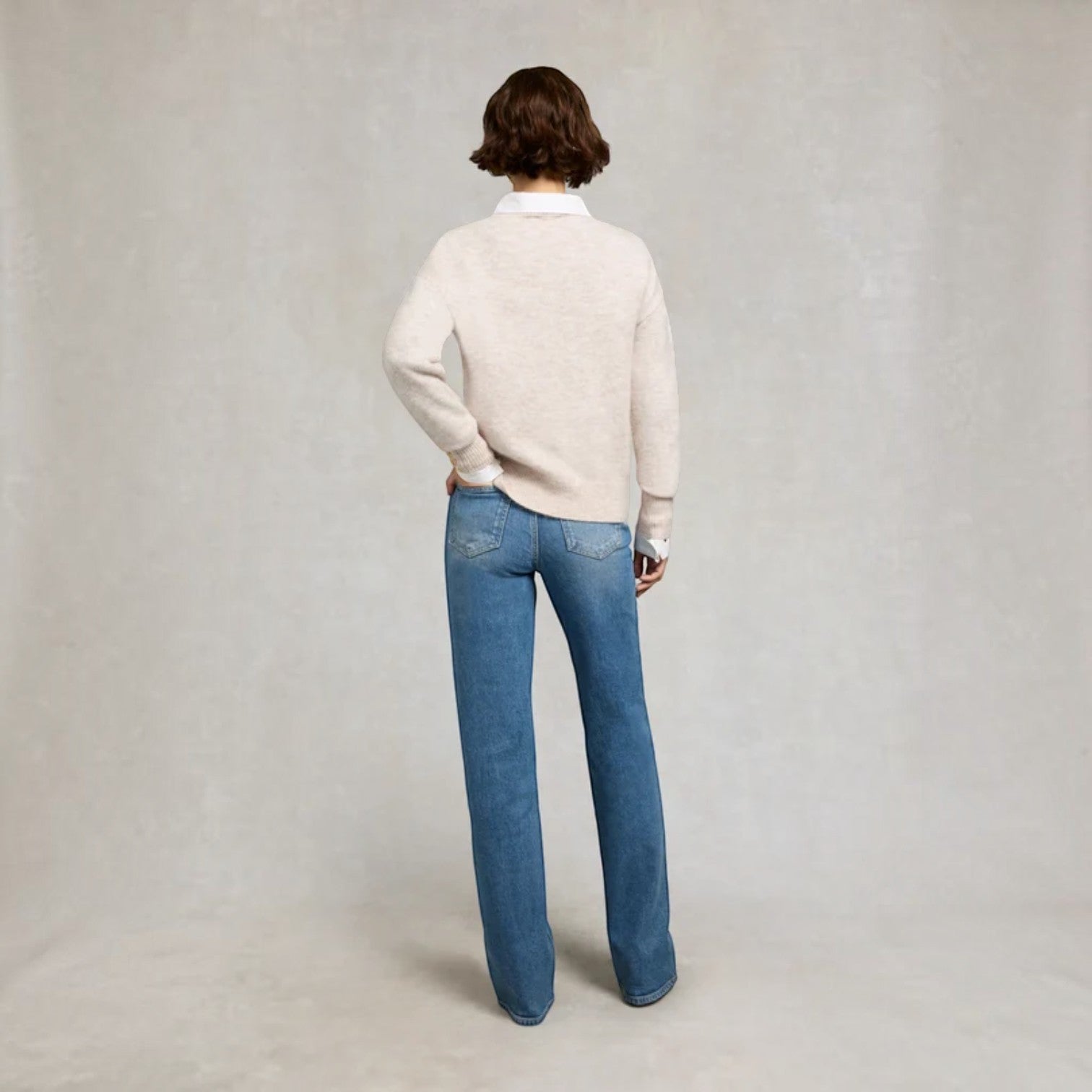 A model viewed from behind wearing a light beige knit sweater over a white collared shirt and blue jeans, standing against a neutral textured gray studio backdrop.