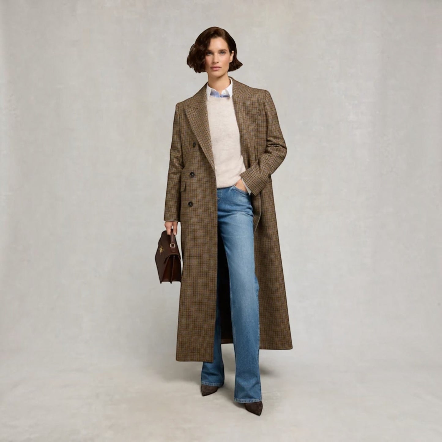 A model stands against a light gray textured studio backdrop wearing a long brown and beige checkered overcoat over a cream sweater, blue jeans, and dark brown pointed shoes, while holding a dark brown handbag.