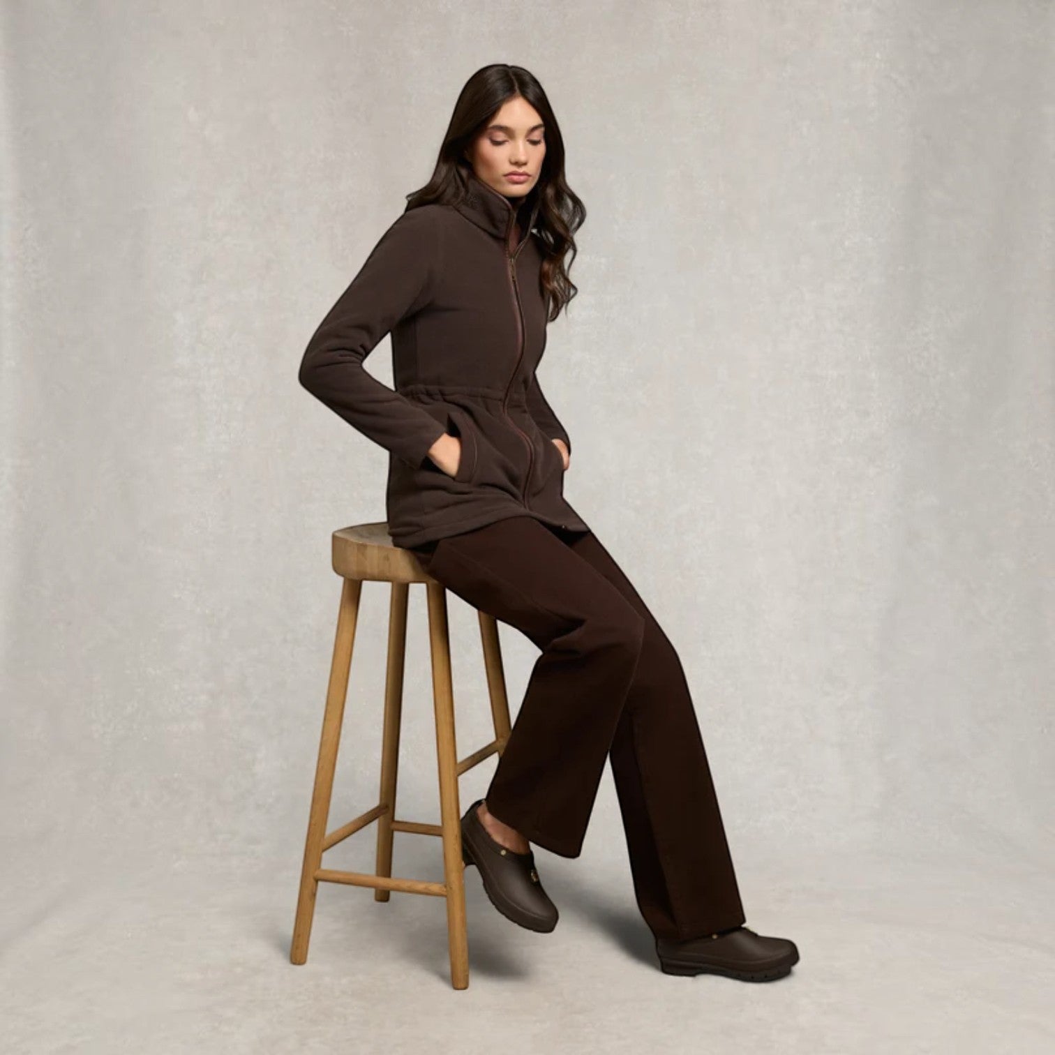 Woman in a brown outfit sitting on a wooden stool against a plain background