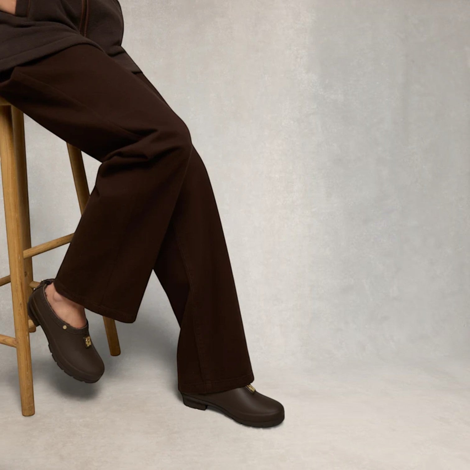 A person seated on a wooden stool wearing dark brown wide-leg trousers and matching dark brown slip-on shoes against a light gray textured studio background.