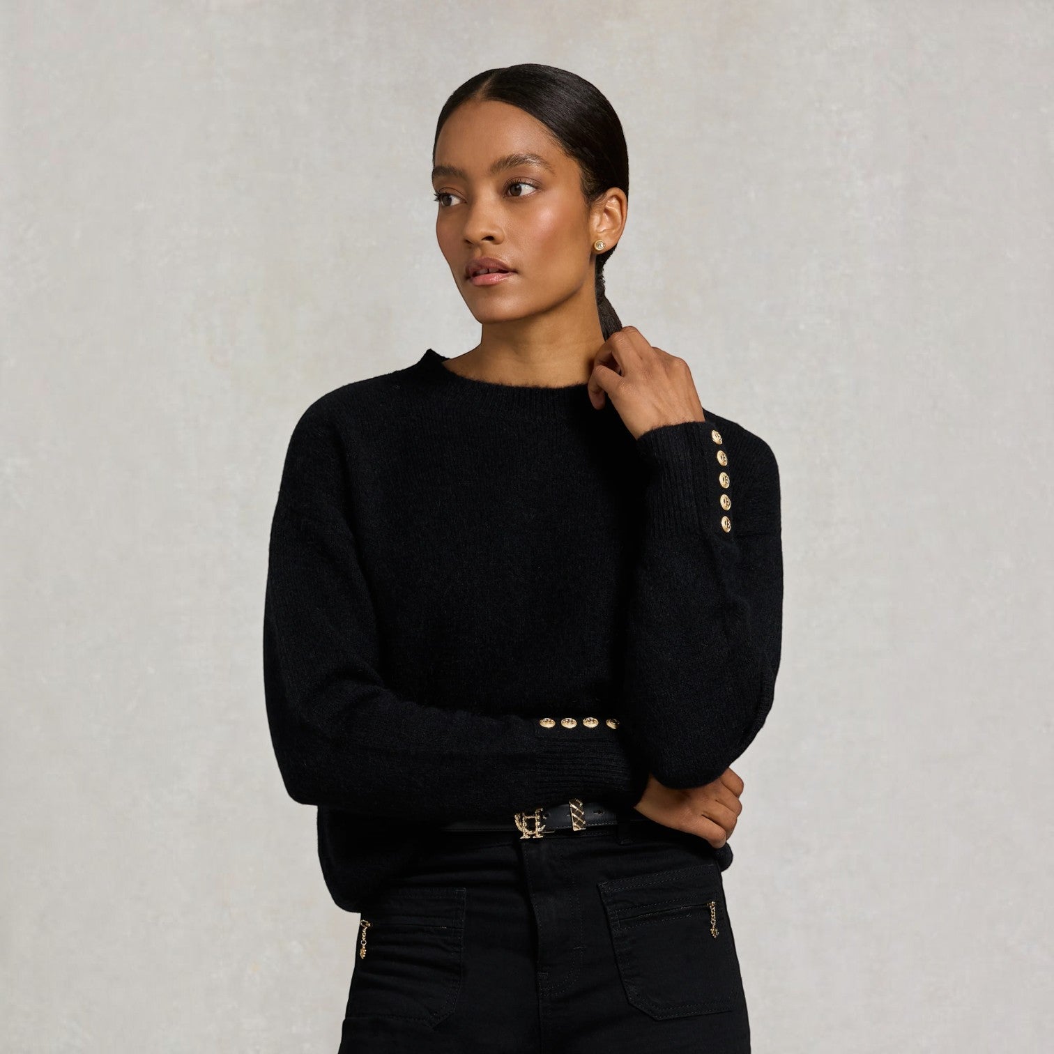 A woman wearing a black knit sweater with decorative gold buttons on the cuffs and waist, paired with black trousers, posed against a textured light gray background.