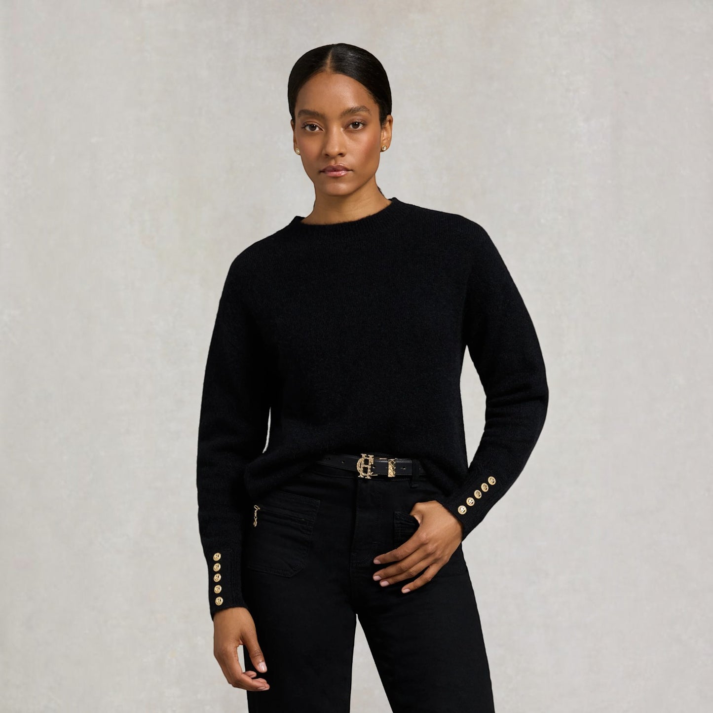 A model wearing a black long-sleeve knit sweater with decorative gold buttons on the cuffs, paired with black jeans and a gold belt, against a textured light gray background.