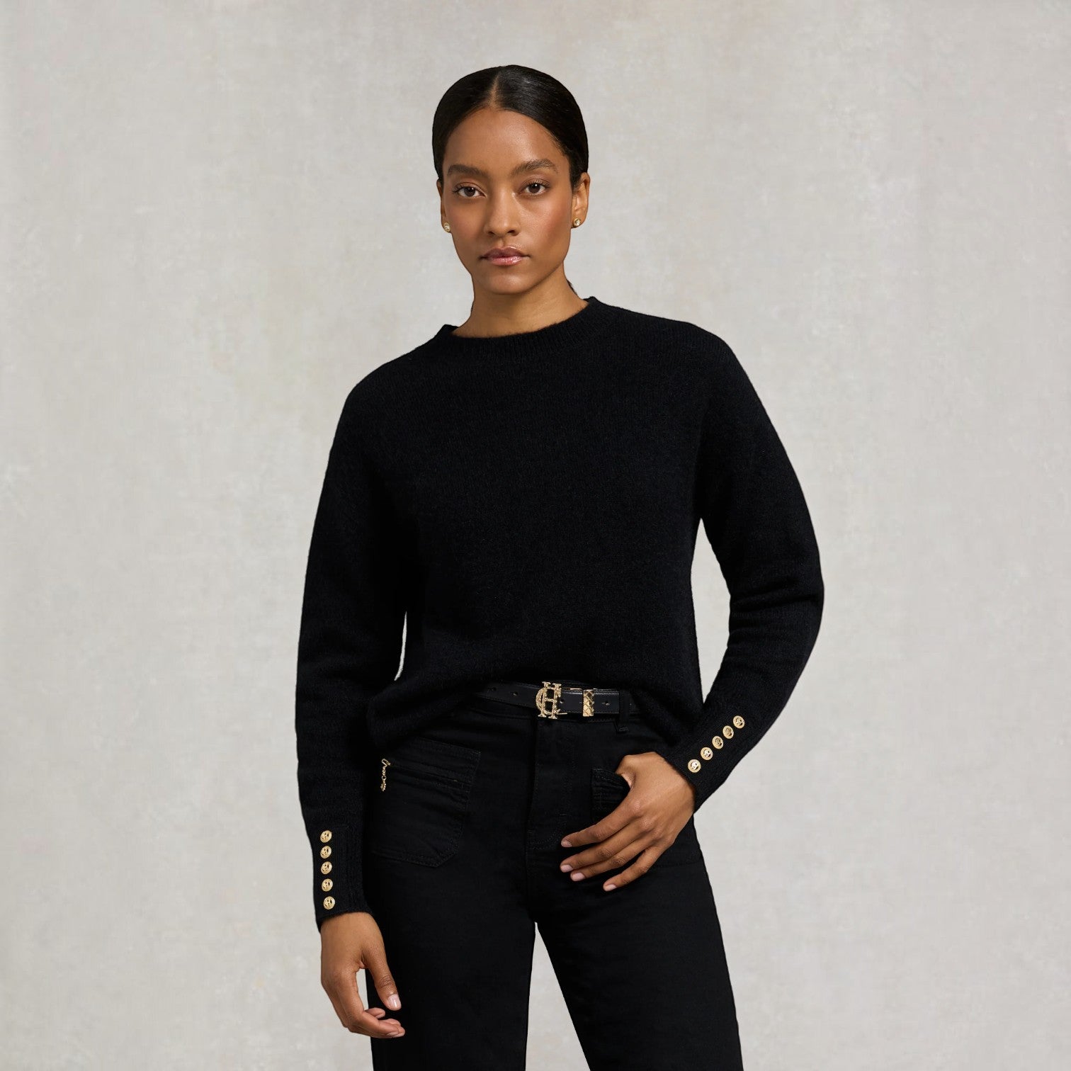 A model wearing a black long-sleeve knit sweater with decorative gold buttons on the cuffs, paired with black jeans and a gold belt, against a textured light gray background.