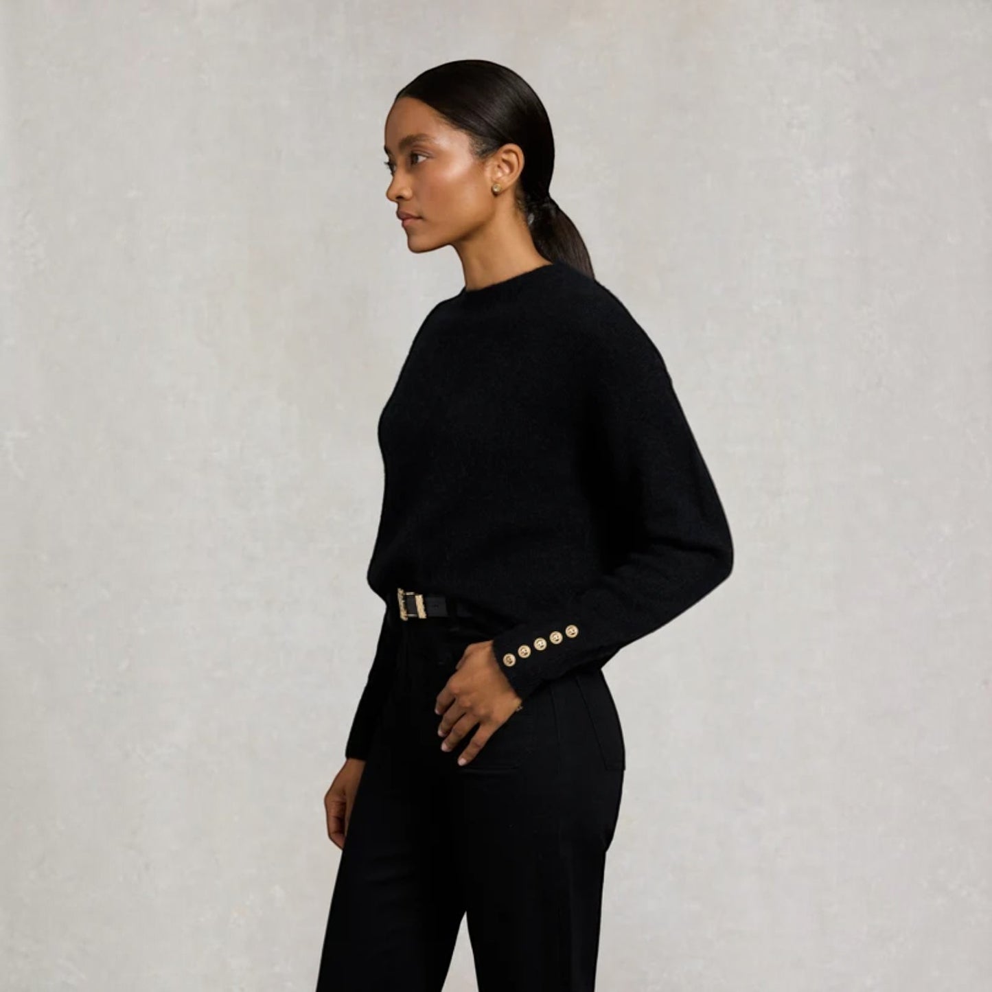 A woman in a black long-sleeve knit sweater and black trousers, shown in profile against a textured light gray wall. The sweater has decorative gold buttons on the cuffs.