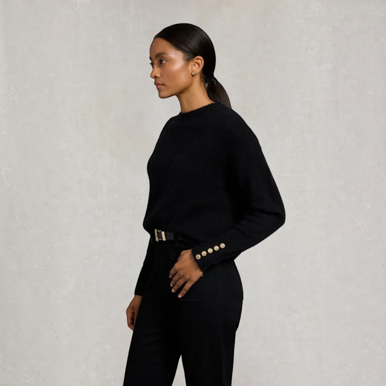 A woman in a black long-sleeve knit sweater and black trousers, shown in profile against a textured light gray wall. The sweater has decorative gold buttons on the cuffs.