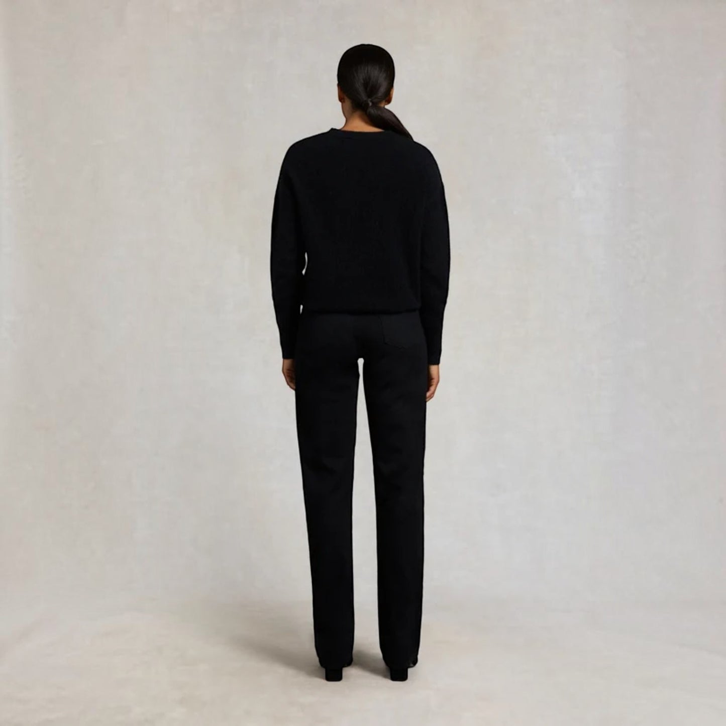 A model viewed from the back wearing a black knit sweater and matching black trousers, standing against a neutral light gray textured studio backdrop.