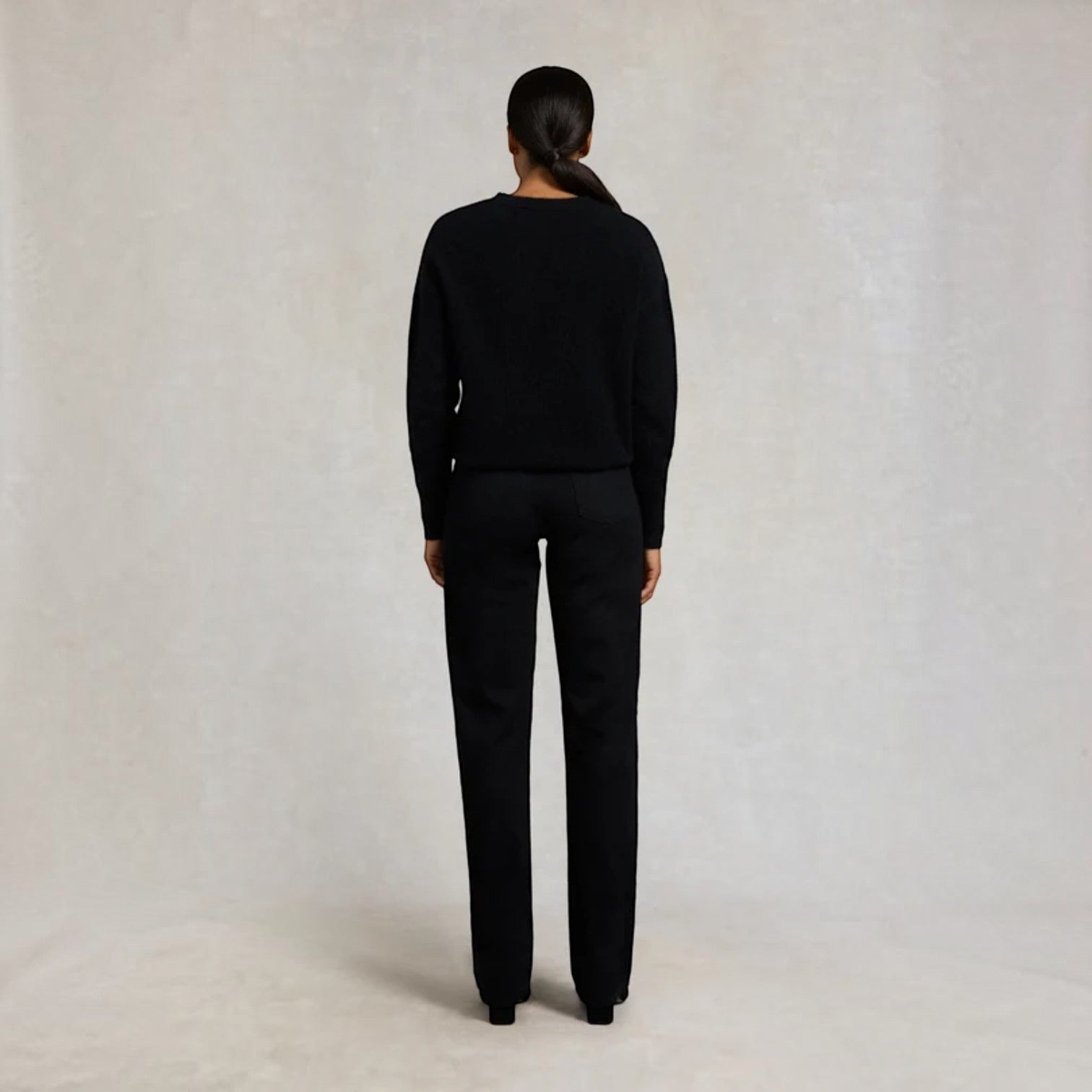 A model viewed from the back wearing a black knit sweater and matching black trousers, standing against a neutral light gray textured studio backdrop.