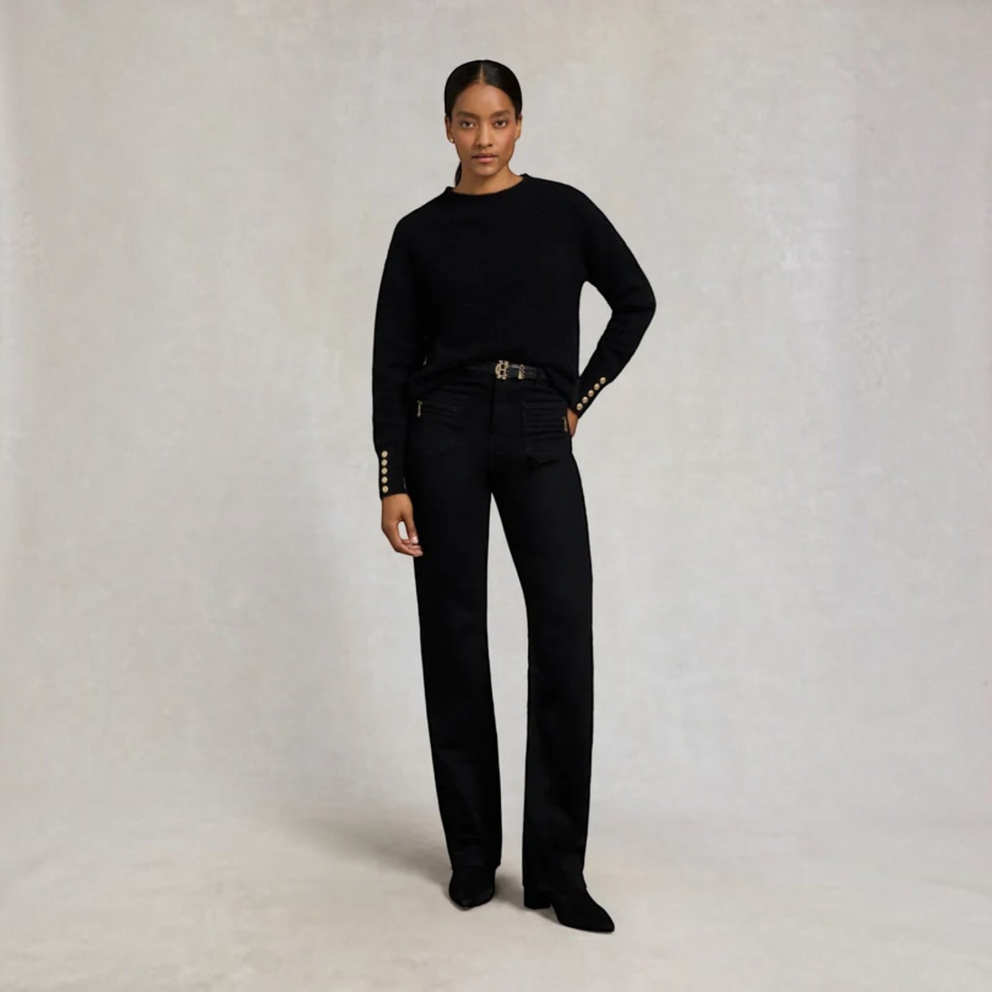 A model stands against a textured light gray studio backdrop wearing a black long-sleeve knit top with decorative gold-tone studs on the cuffs, black high-waisted straight-leg jeans with a gold-tone belt, and black pointed-toe heels.