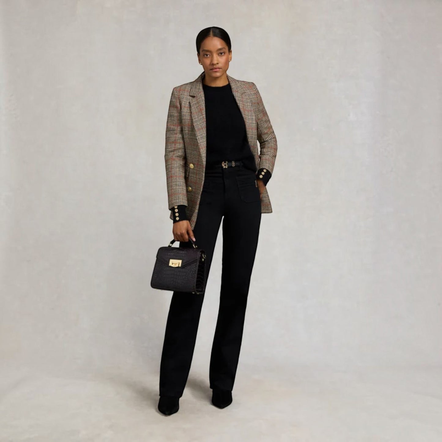 A model stands against a textured light gray studio backdrop wearing a brown and beige plaid blazer over a black top and black trousers, holding a black quilted handbag with gold hardware.