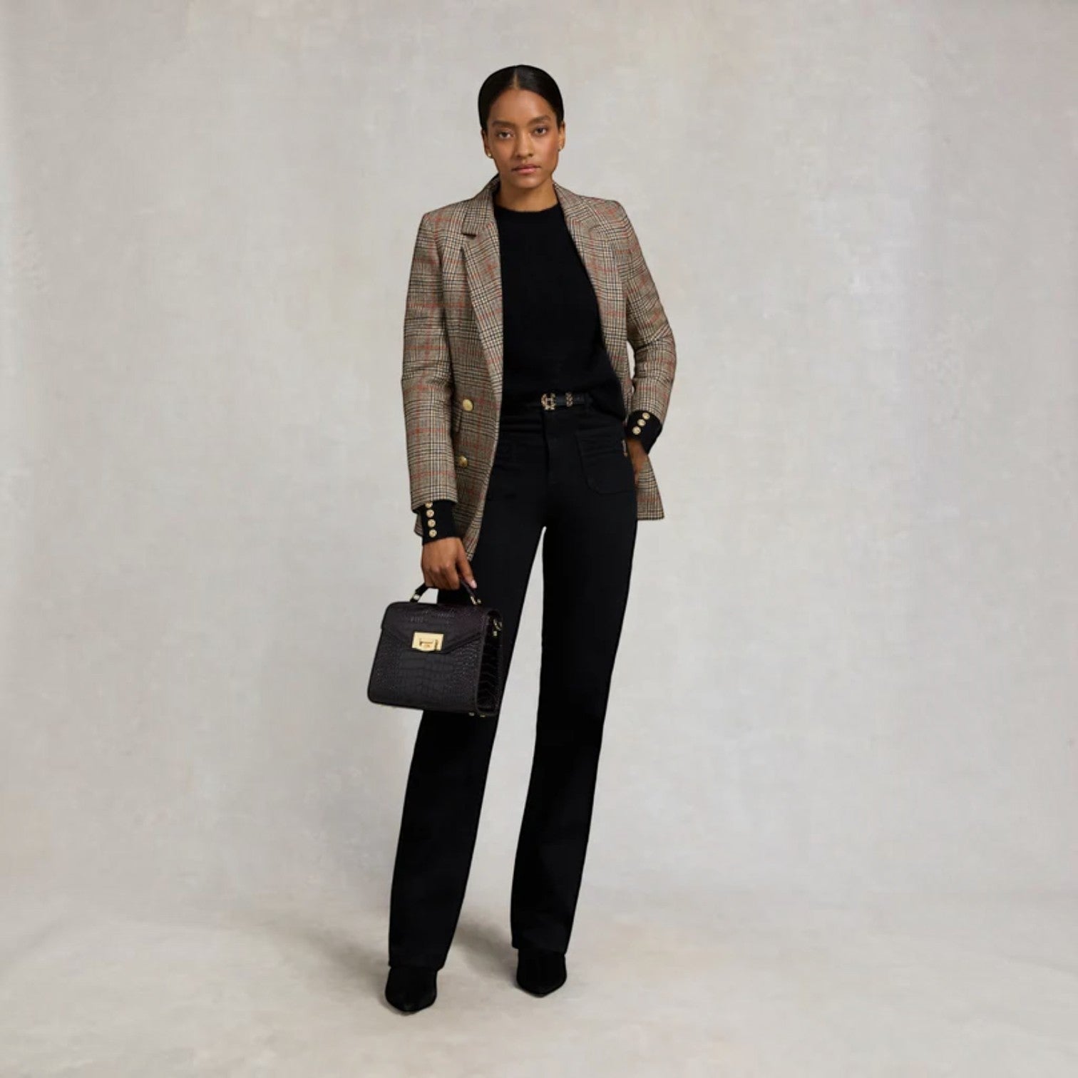 A model stands against a textured light gray studio backdrop wearing a brown and beige plaid blazer over a black top and black trousers, holding a black quilted handbag with gold hardware.