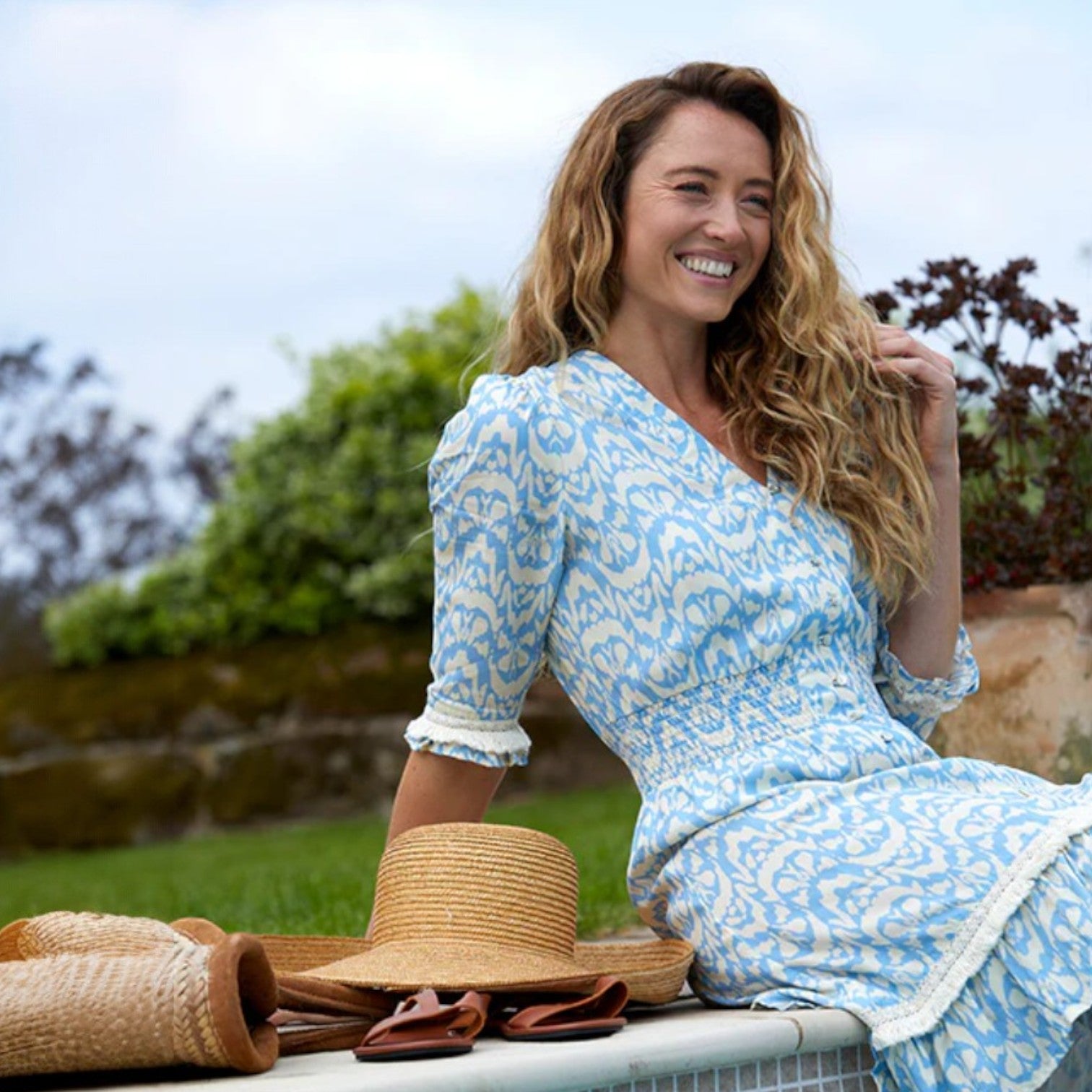 Woman in a blue floral dress sitting outdoors with hats on a table in front of her.