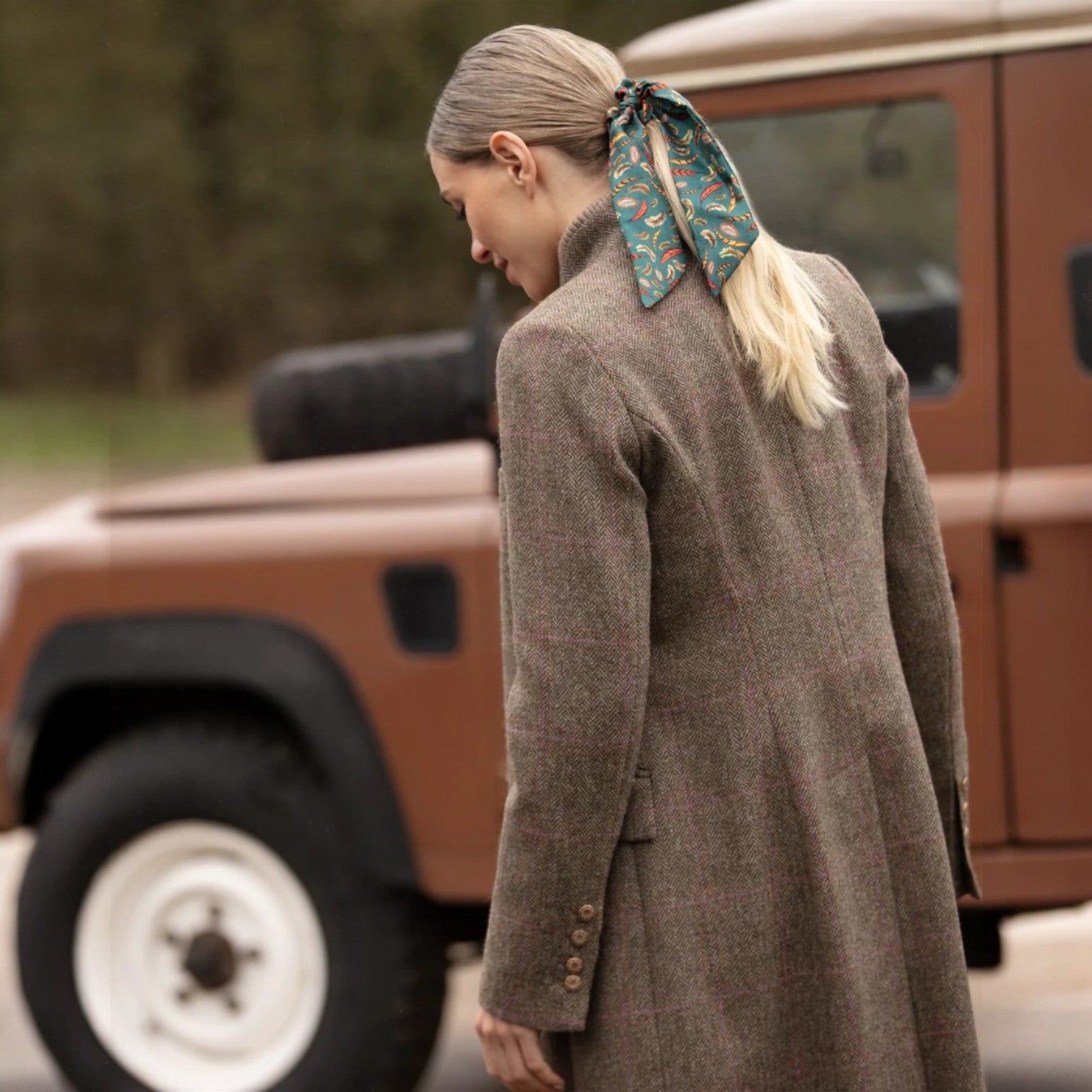 Woman in a brown coat with a patterned scarf in front of a brown vehicle