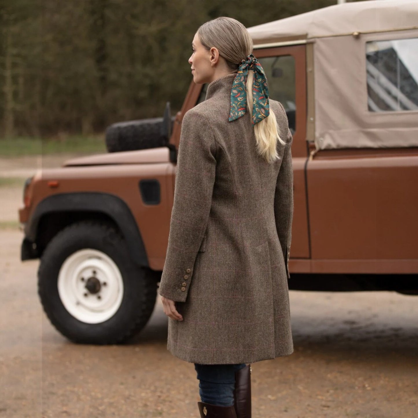 Woman in a brown coat standing next to a vintage brown vehicle in a natural setting