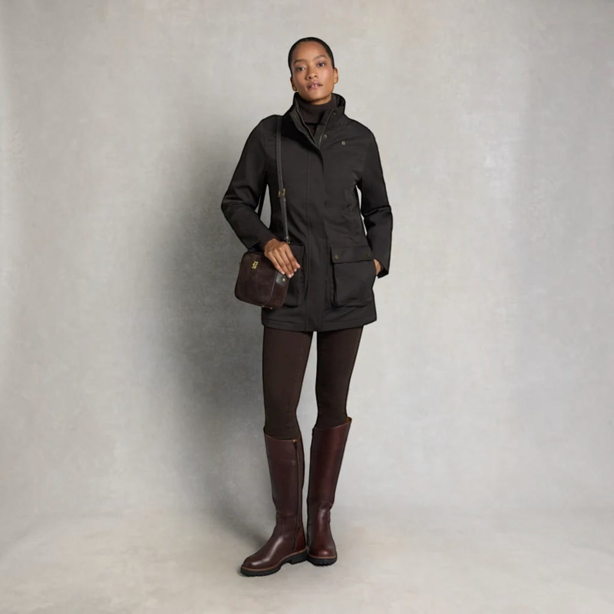 A model stands against a textured gray studio backdrop wearing a black mid-length jacket over a black turtleneck, dark brown leggings, and dark brown knee-high boots. She carries a dark brown leather shoulder bag.