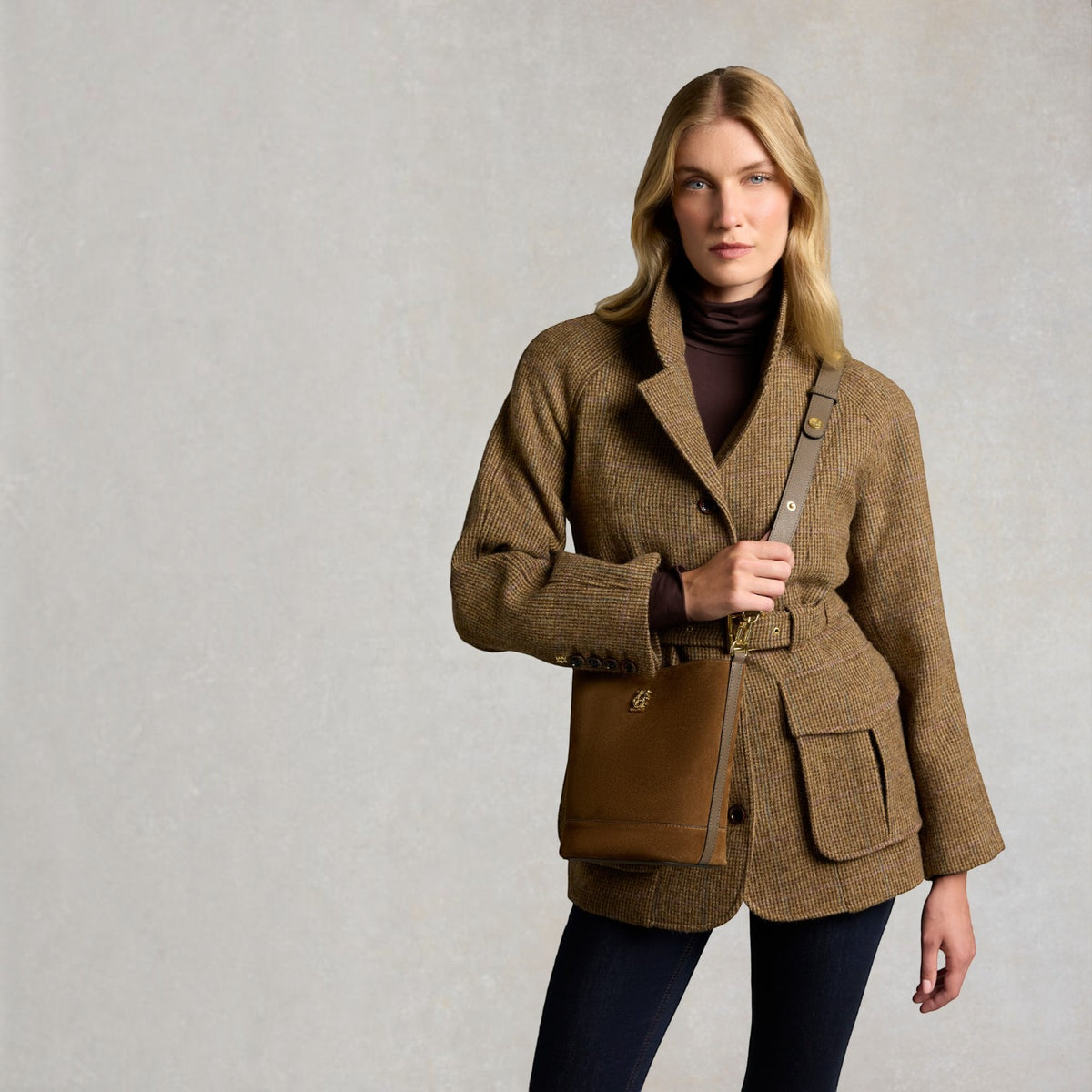 A woman models a brown tweed belted jacket with a brown leather crossbody bag featuring a gold 'Burberrys' monogram, set against a textured light gray studio background.